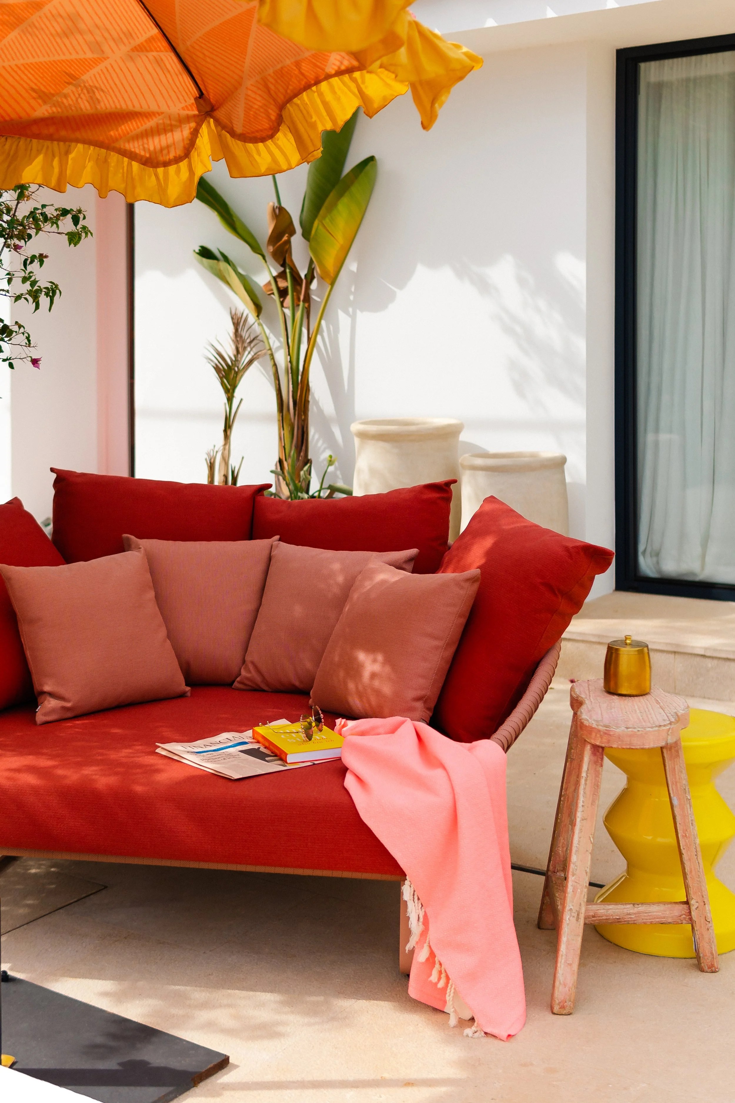 Outdoor patio scene featuring a pink and red cushioned lounge sofa with several pillows, pink blanket, magazines, a small yellow table, a weathered pink wooden side table with a small gold container, a large yellow and orange patio umbrella, tall green plants, and a sliding glass door.
