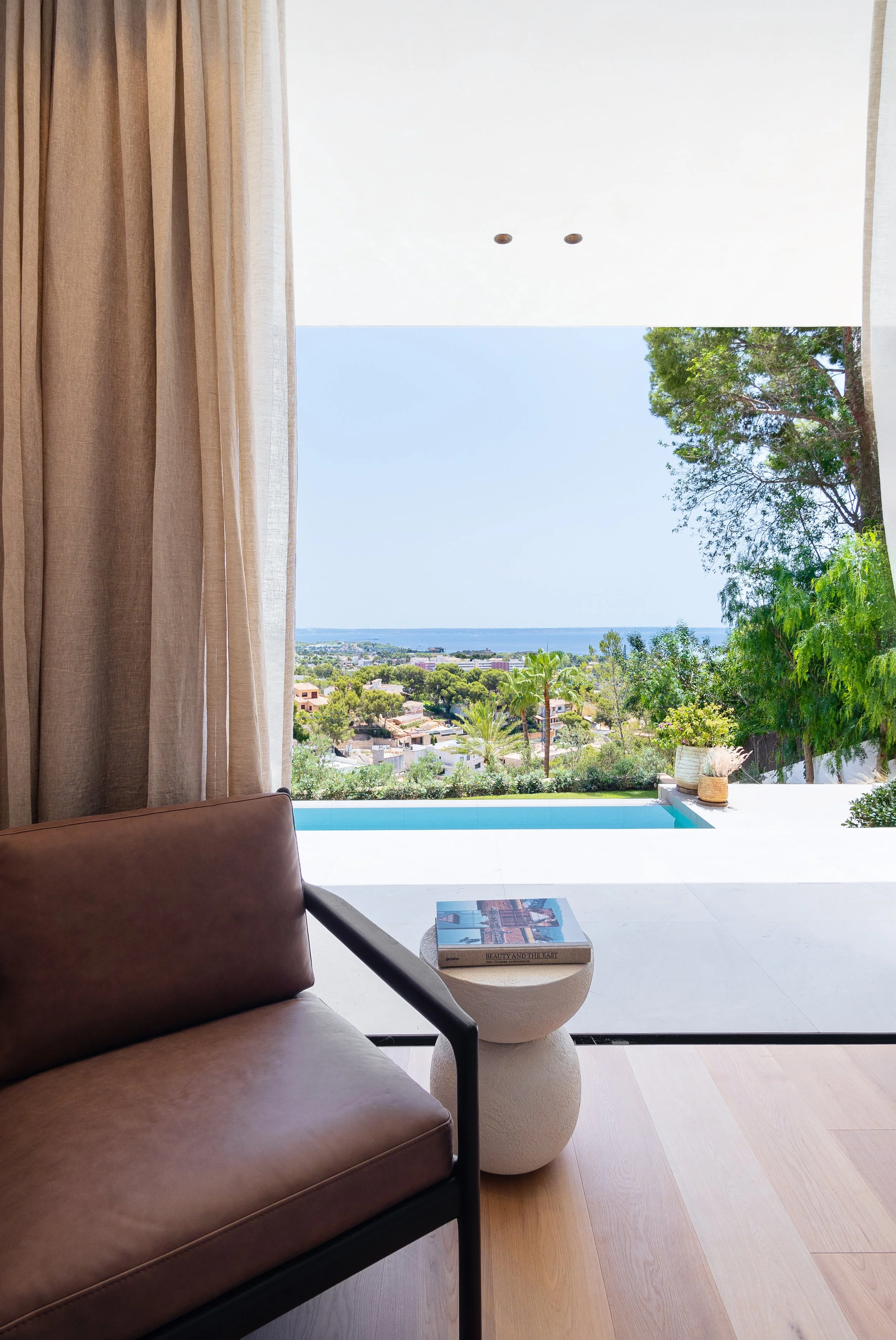 Living room with glass door showing view of trees, houses, and ocean, brown armchair, dark side table with a book, beige curtains, wooden floor, and potted plants outside.