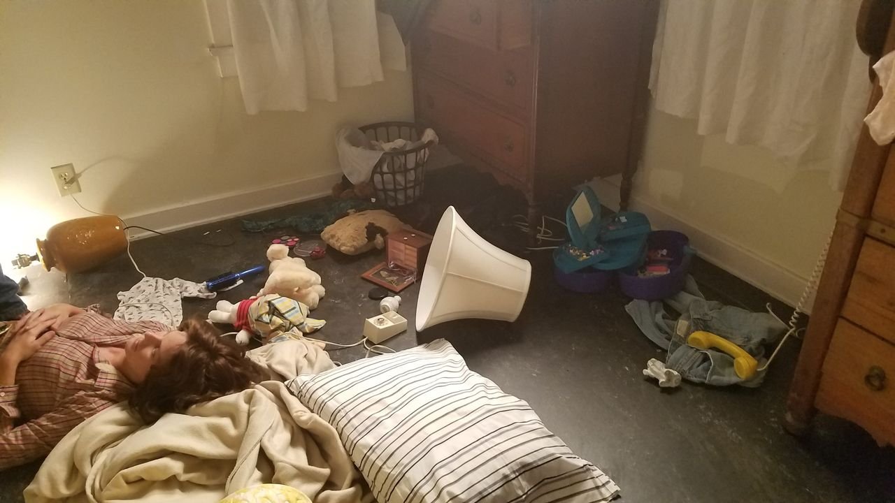 A bedroom with a pile of stuffed animals, a broken lamp, a fallen paper lantern, a fallen lampshade, and scattered items including a phone, boxes, and clothes. Two people are lying on the floor among the clutter. Scene from 'The Innocent Man'.