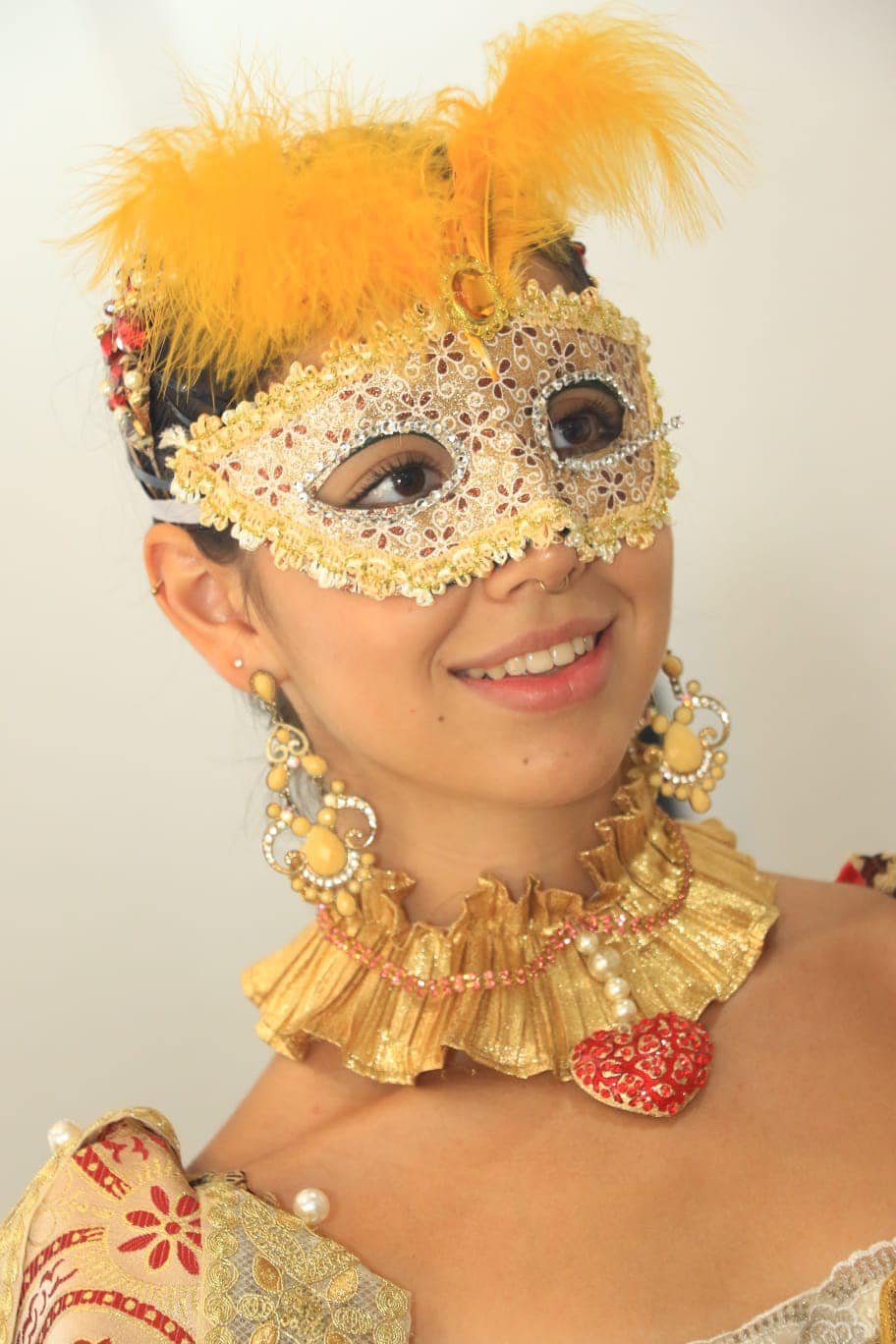 Juliet Capulet actress wearing a decorative yellow gold and red Venetian-style mask with feathers, earrings, and a ruffled gold collar, smiling with a pearl necklace featuring a red heart pendant.