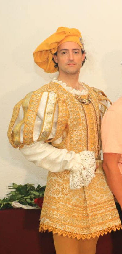 Romeo Montague actor dressed in a Renaissance-style costume with ornate gold and white fabric, a padded sleeve, a ruffled collar, and a yellow hat, standing against a cream-colored wall.