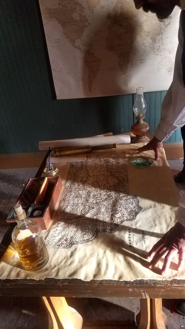 An old map spread out on a wooden table, with a person pointing at it. There is a bottle of whiskey, a quill pen, a glass oil lantern, an inkwell, and rolled-up scrolls on the table. A large world map hangs on the wall behind. Props by Lauren Marina.