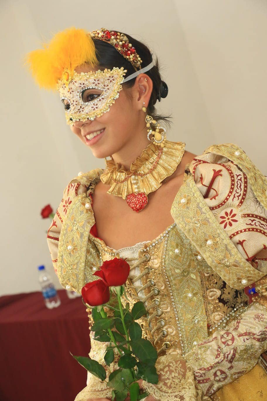 Juliet Capulet actress wearing an ornate gold and red Renaissance-style costume with a lace and pearl mask, holding red roses, smiling.