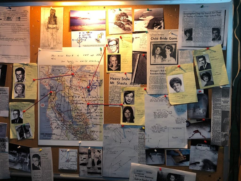 A bulletin board covered with maps, photographs, newspaper articles, notes, and research materials, including images of people, handwritten notes, and pinned papers, indicating an investigation or case study. From 'The Most Dangerous Animal of All'.