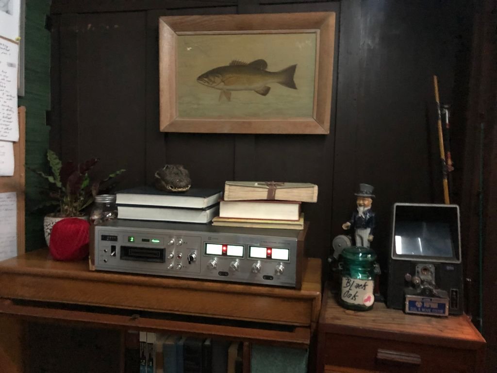 A wooden table with a vintage stereo/tape deck, three stacked books, a frog figurine, a potted plant, and a jar labeled 'Black Oak'. Set dressing from ' The Most Dangerous Animal of All' on FX.