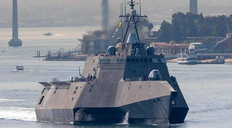 A large gray military warship sailing on a river with bridges, smoke, and smaller boats in the background.