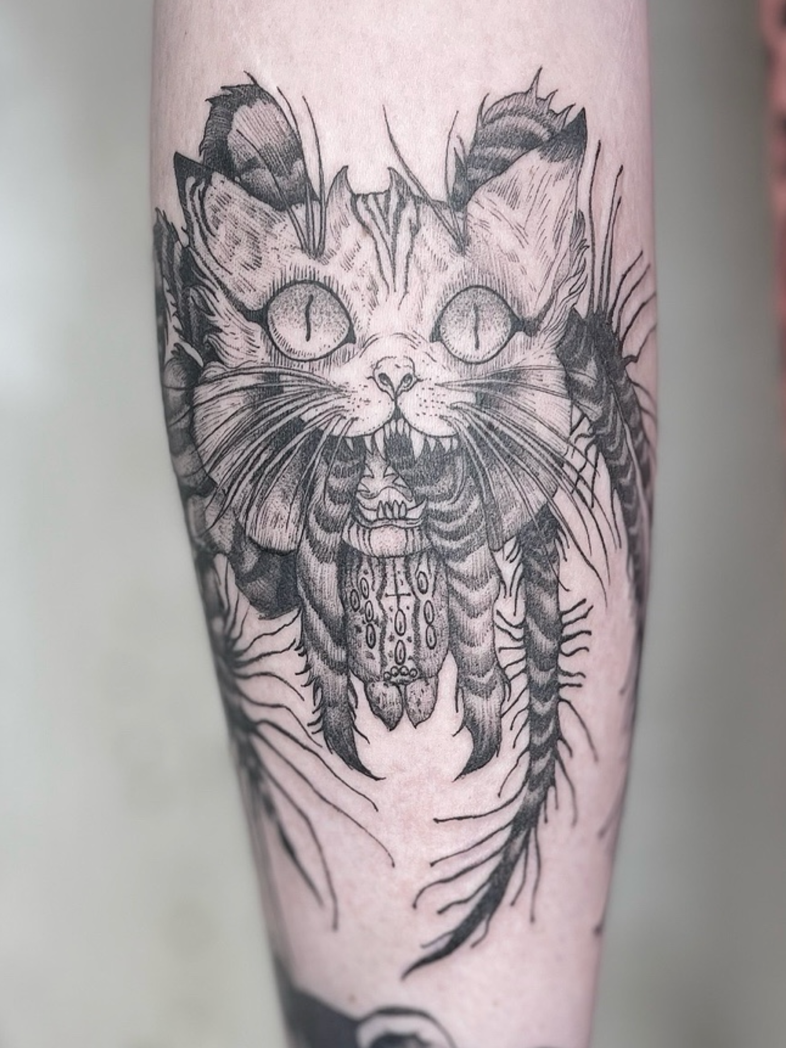 A black and gray tattoo of a ferocious cat with open mouth and spider legs, sharp teeth, and piercing eyes on a person's leg.
