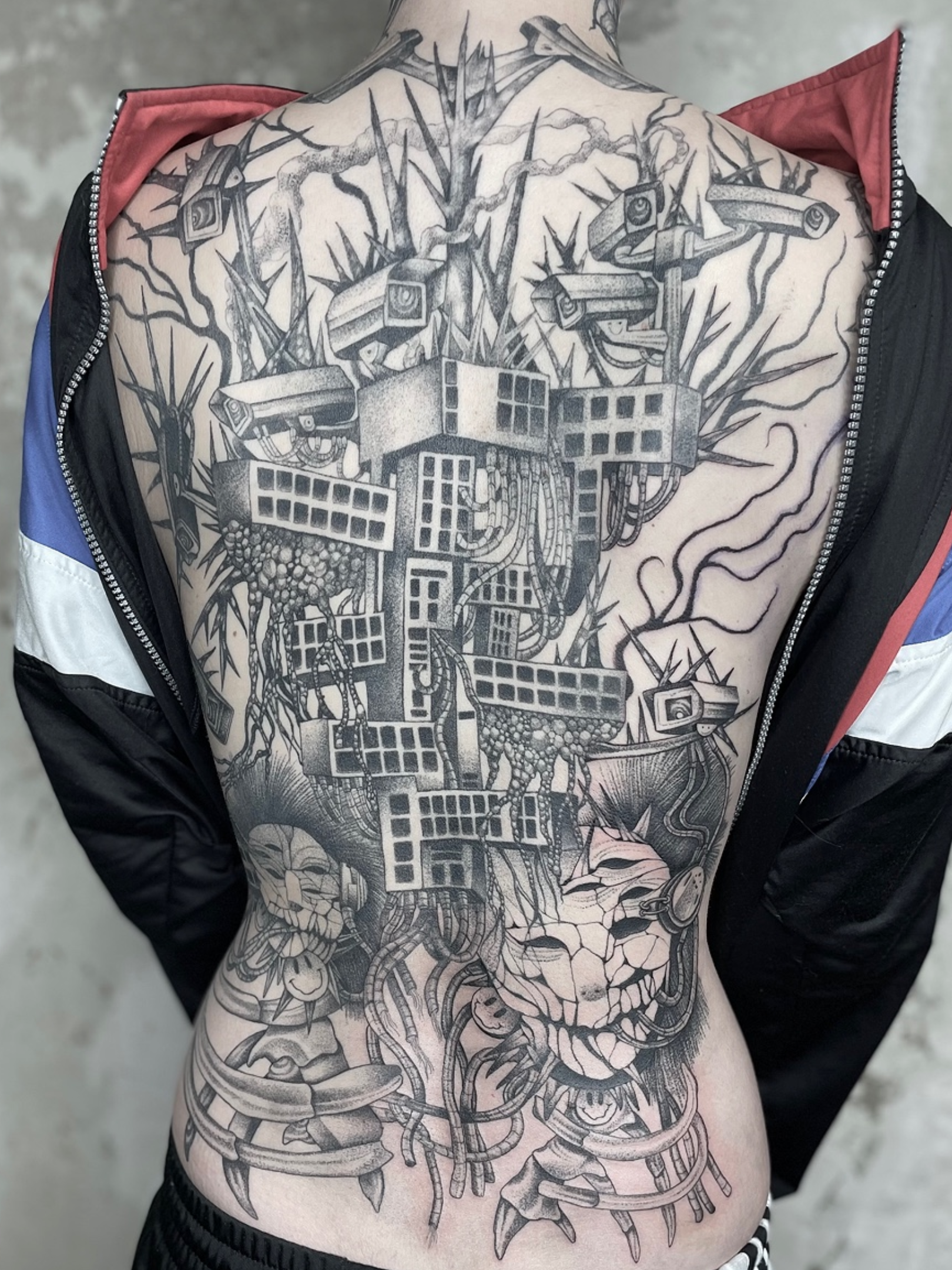 A person's back with a large black and gray tattoo depicting a detailed skull at the bottom and a futuristic cityscape with tall grid-like buildings and multiple surveillance cameras above, with stormy clouds and lightning in the background.