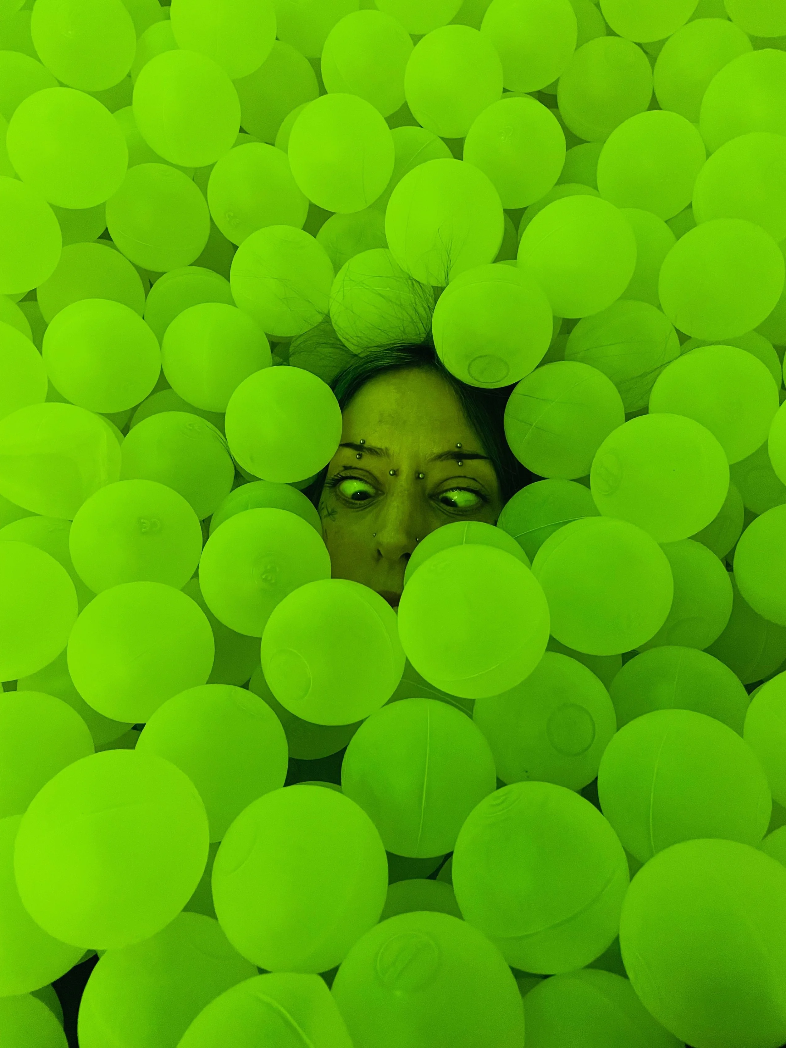 A person with piercings on their face lies among a large number of bright green plastic balls, with only part of their face visible.