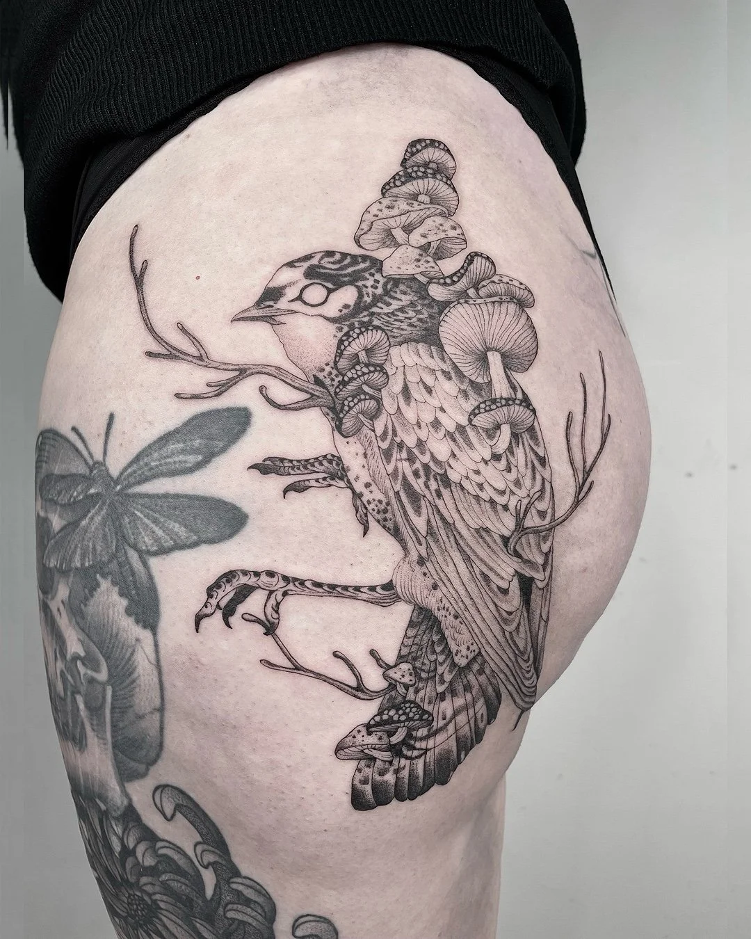 A black and gray tattoo of a bird with mushrooms growing on its back, perched on a branch, with a butterfly nearby on the arm.