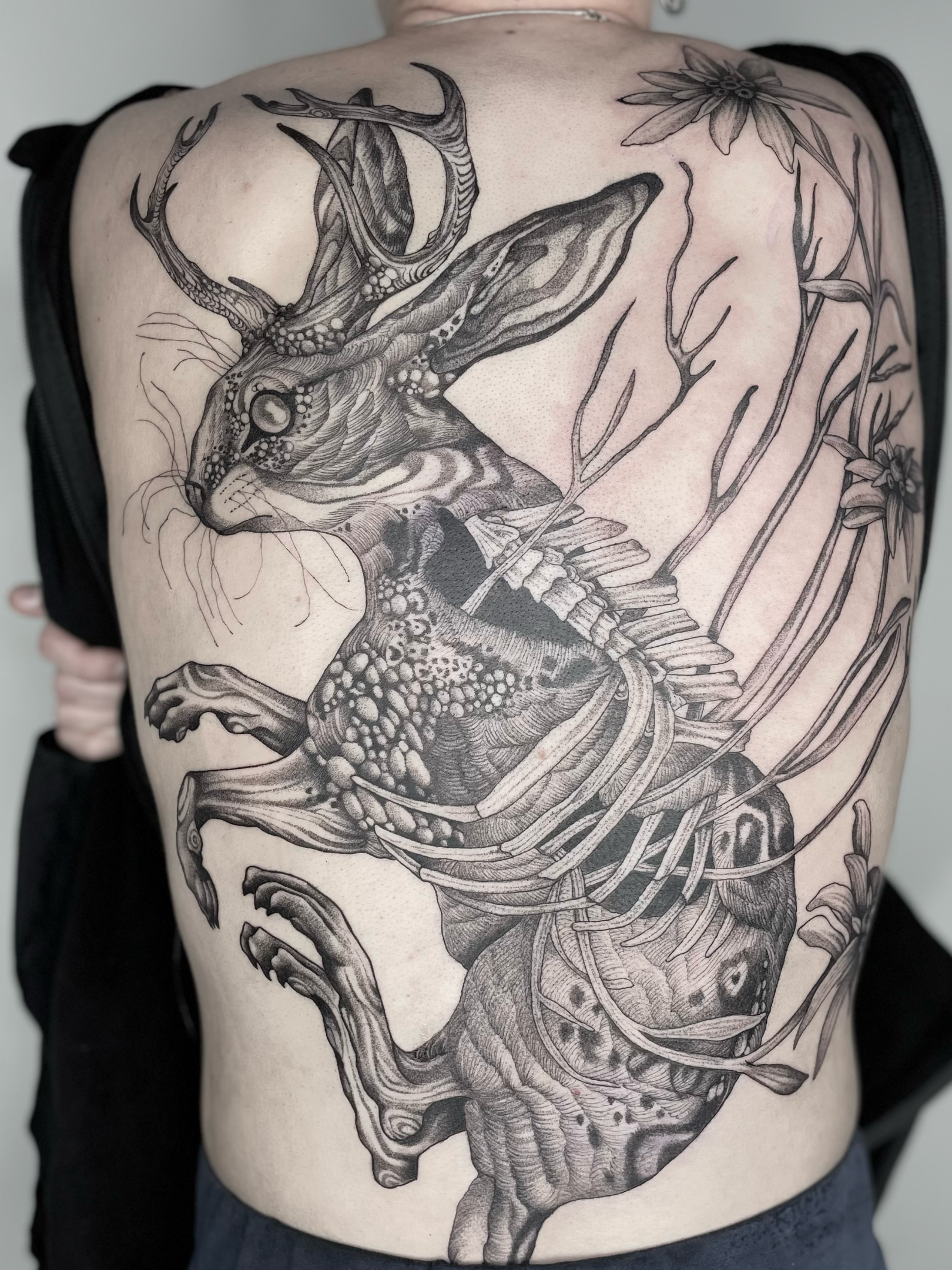 A detailed black and gray tattoo of a rabbit with antlers, surrounded by flowers and plant elements, covering the person's back.