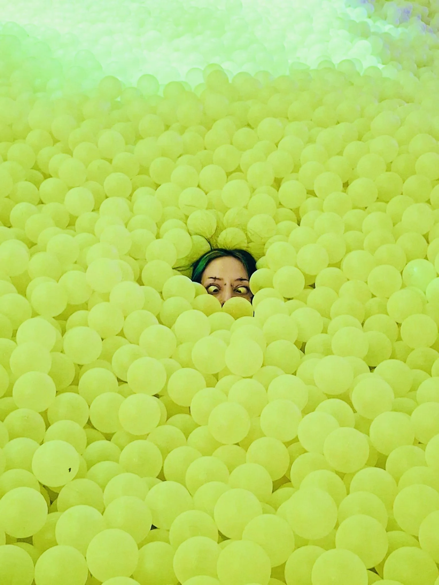 Person partially submerged in a ball pit filled with yellow balls, with only their face visible.