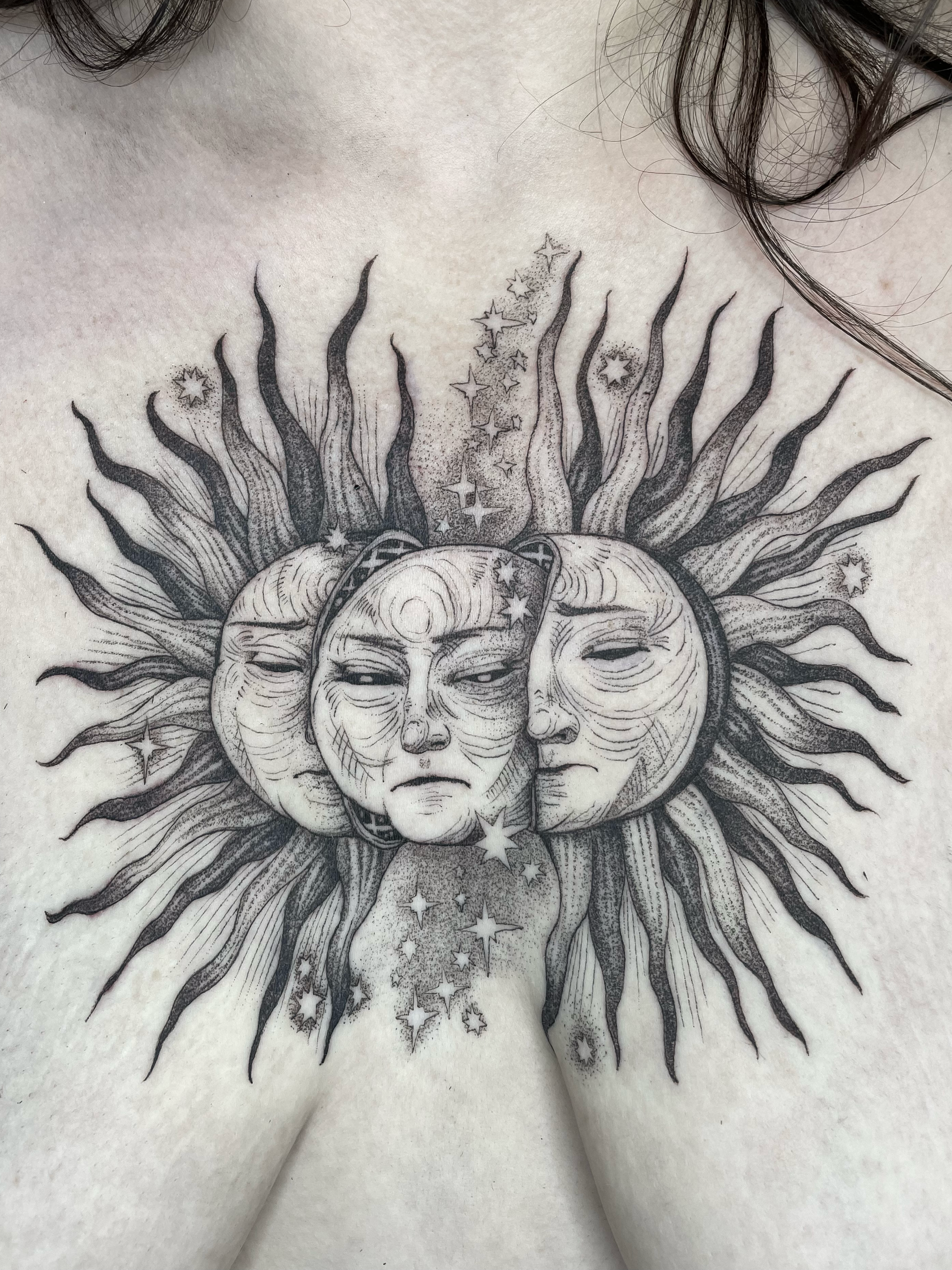 Tattoo of a sun with a serene face, surrounded by radiating rays, stars, and celestial patterns on a person's body.