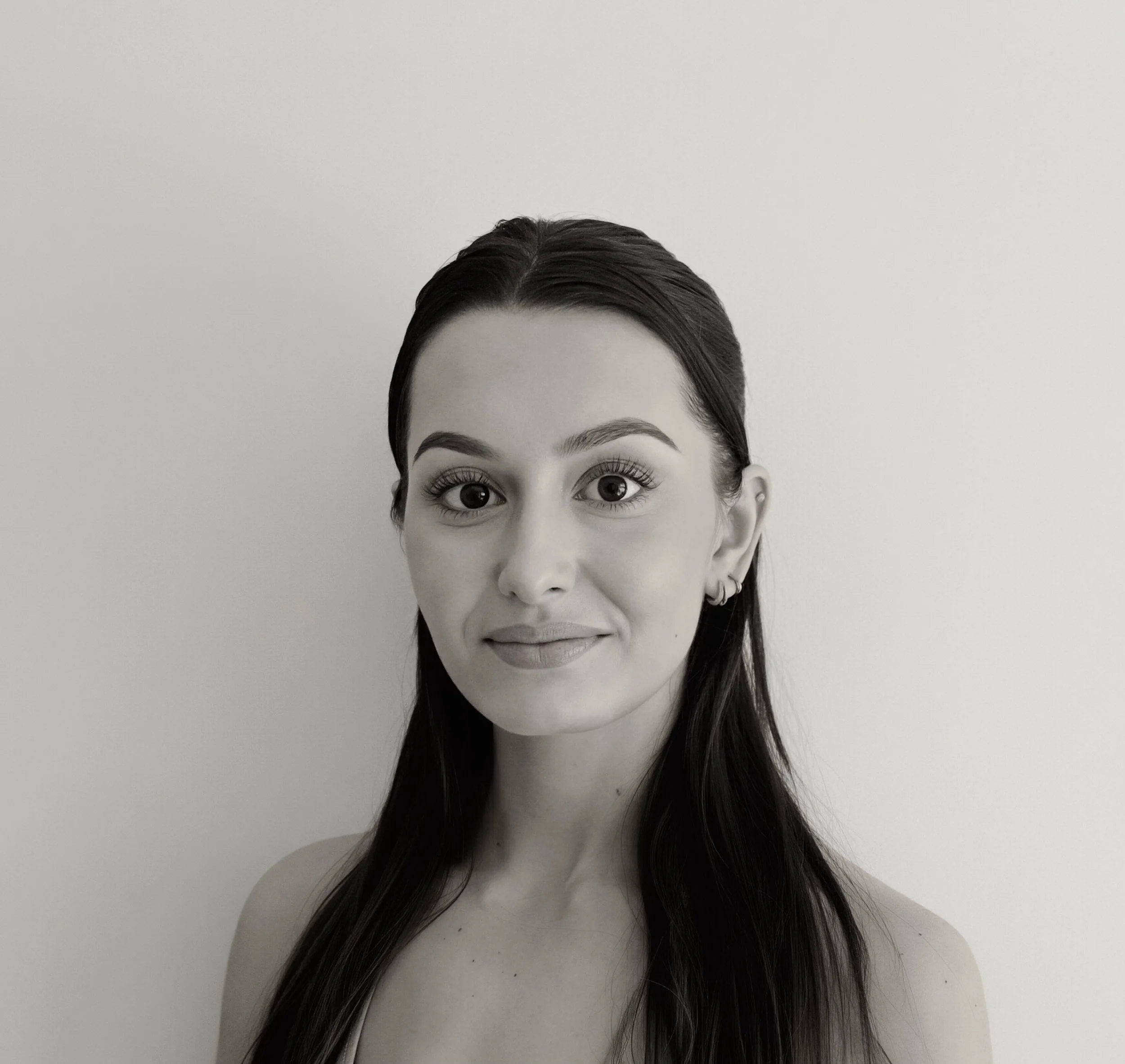 Black and white portrait of a young woman with hair tied back, wearing a zip-up athletic jacket with a high collar and hoop earrings, standing against a plain light-colored wall.