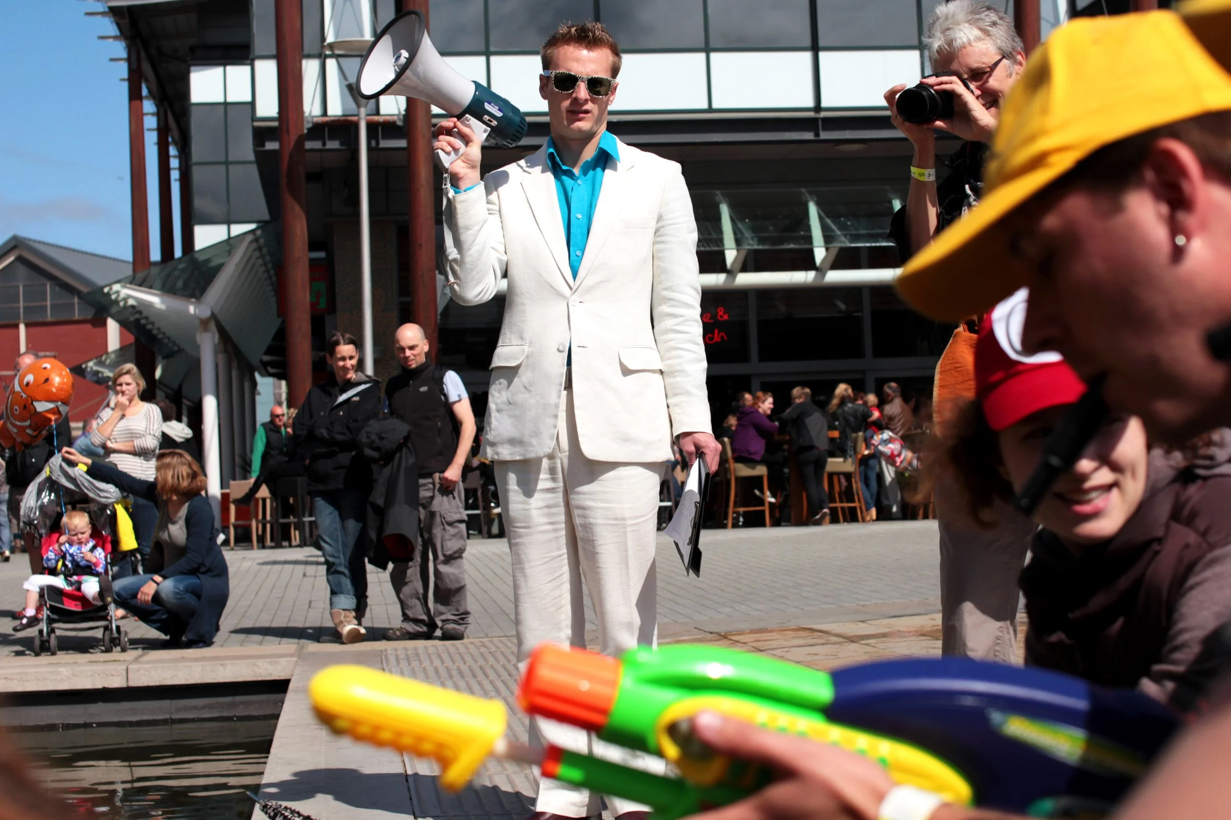 A man in a white suit stands with a loudspeaker, overseeing a water fight in the street.