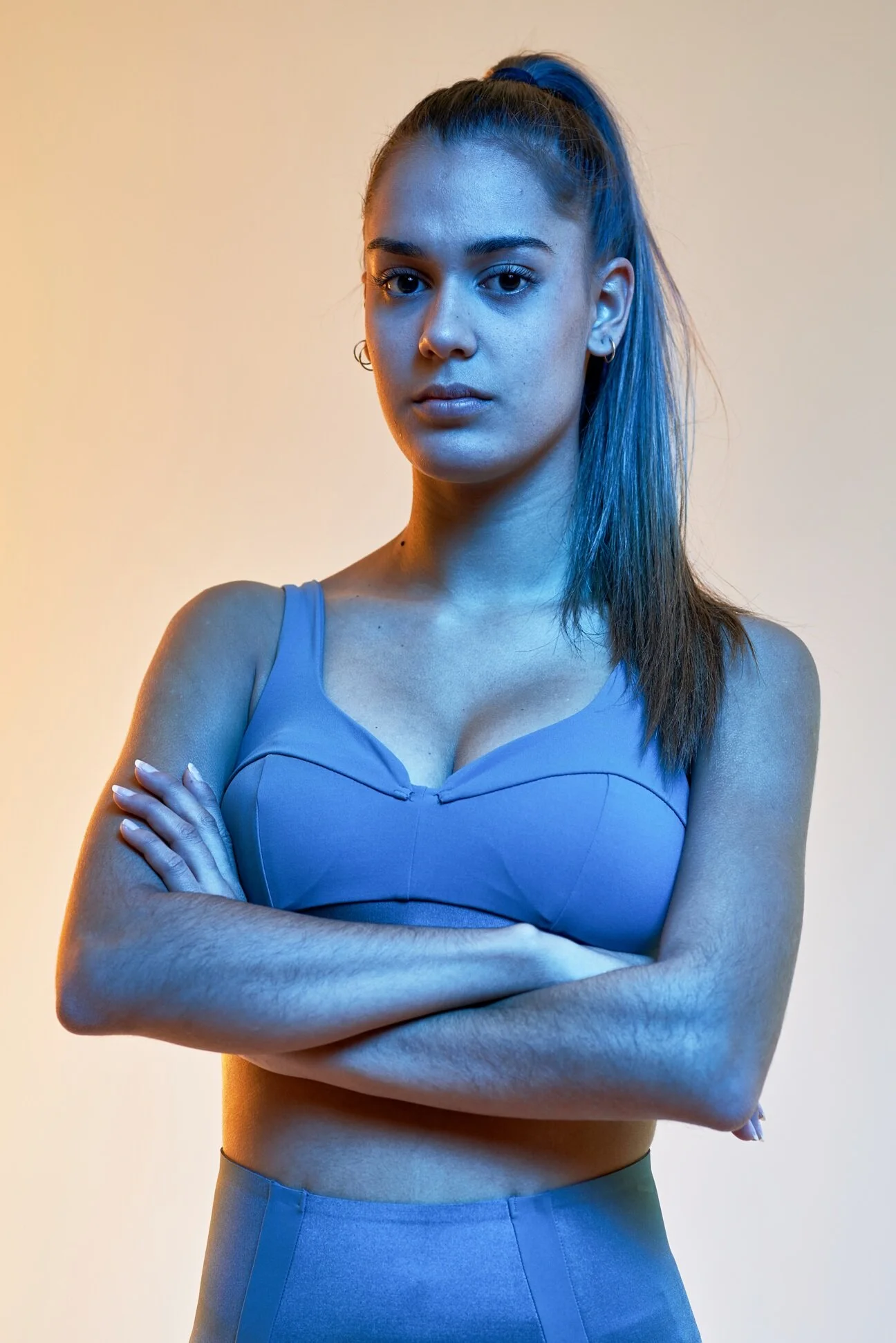 Joven mujer con cabello alarrollado en una cola de caballo, sin expresión y con brazos cruzados. Usa un top azul y pantalones de talle alto.
