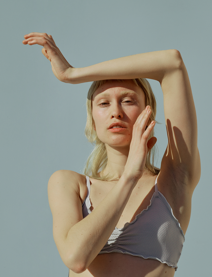 Junge Frau mit blonden Haaren und heller Hautzeit steht im Freien bei Sonnenlicht. Sie trägt ein grau-weiß gestreiftes Top mit dünnen Trägern. Ihre linke Hand berührt ihr Gesicht, während ihre rechte Arm über ihren Kopf gehoben ist.