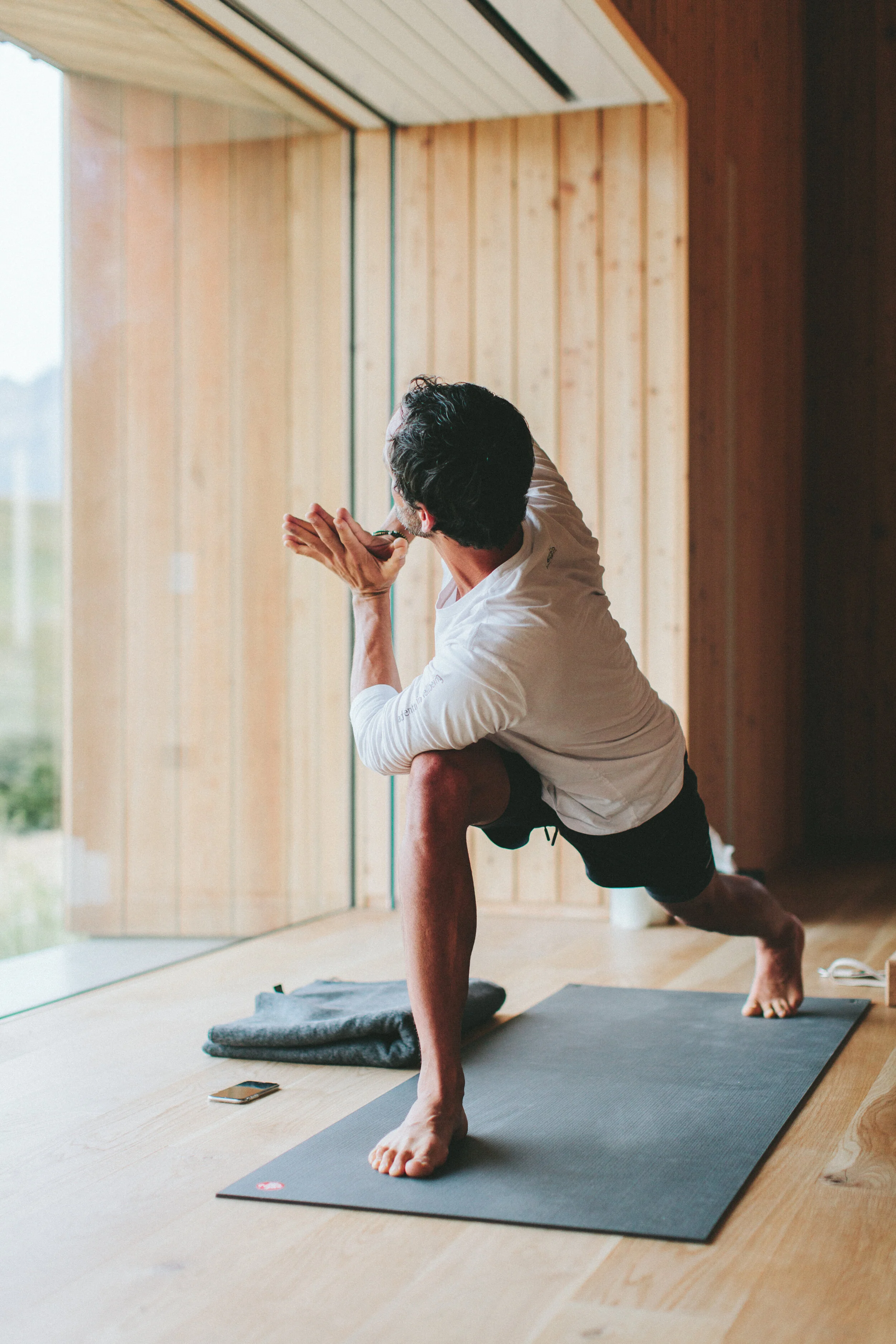 A man doing yoga in a wooden room near large windows, in a lunge pose with hands together in prayer, on a yoga mat.