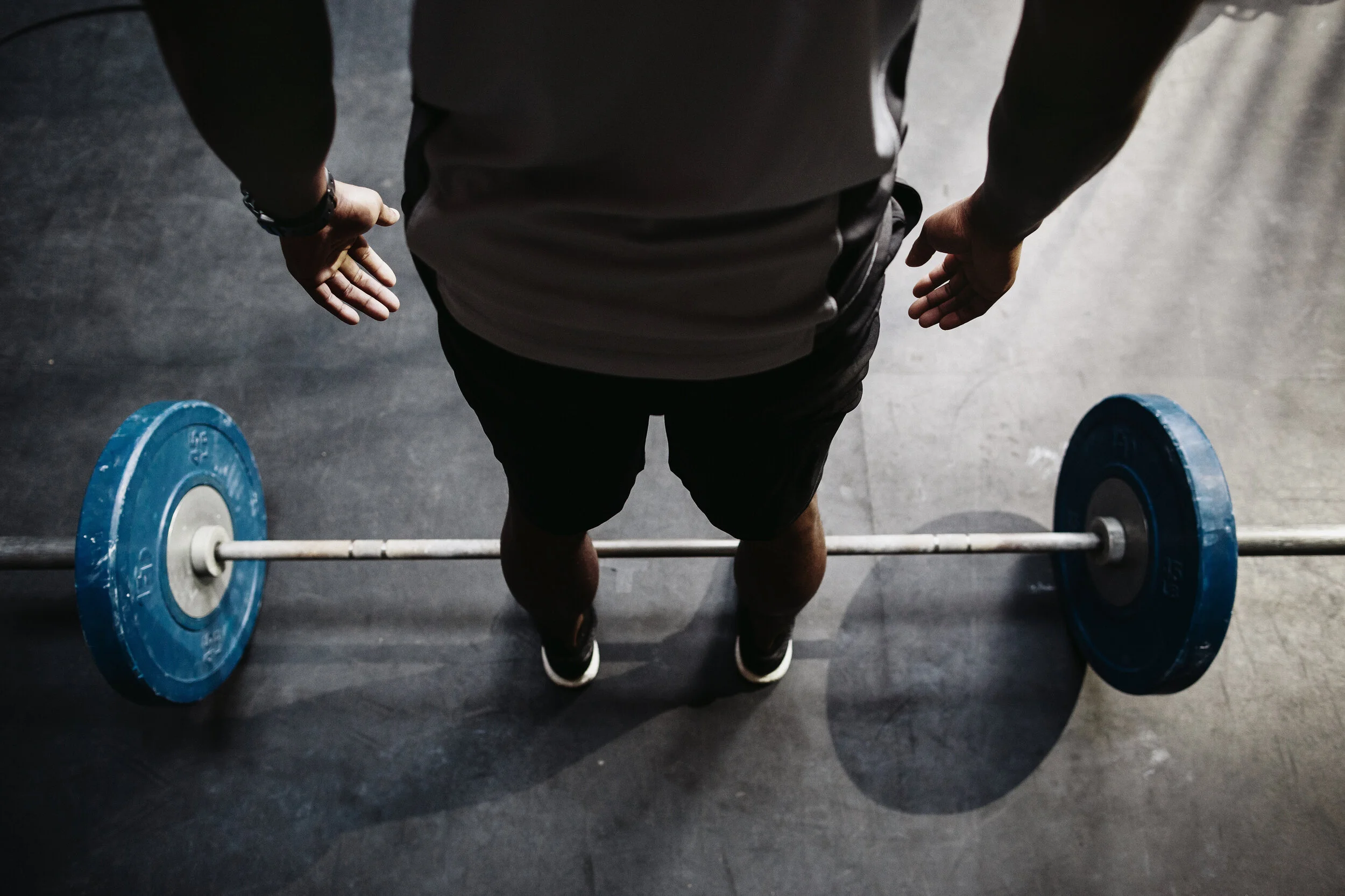 From a top-down view, a man in a dark shirt and shorts stands in a gym next to a barbell loaded with blue weight plates on a black floor, preparing to lift.