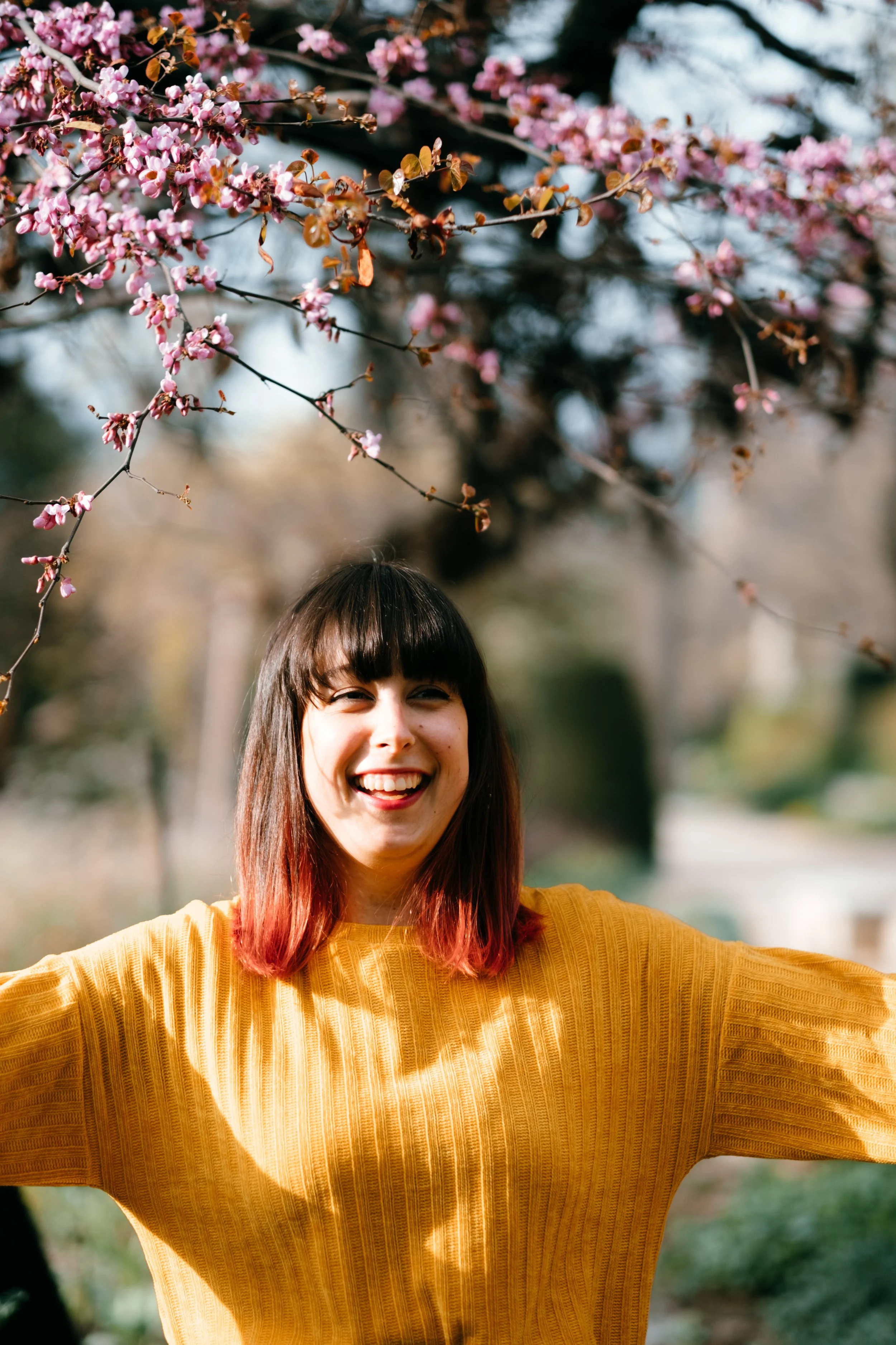 Donna con capelli lunghi fino alle spalle, con balayage dal castano scuro al rosso brillante. Sorride circondata da fiori di ciliegio.