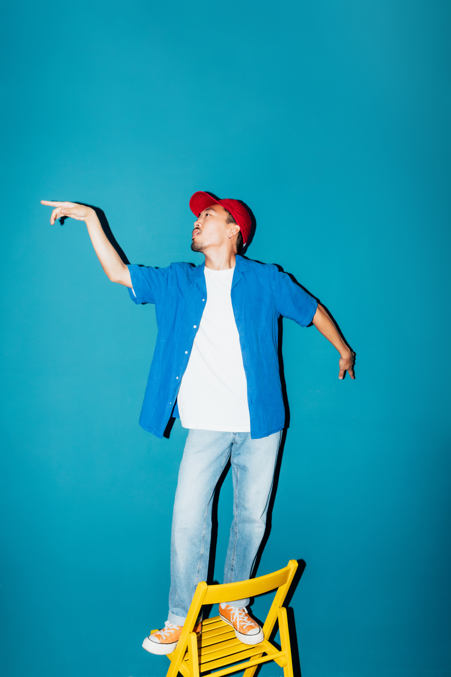 Giovane uomo asiatico ritratto in una posa stravagante, in piedi su una sedia pieghevole gialla. Guarda in lontananza e indica con un braccio. Lo scatto è ambientato su uno sfondo color verde acqua. Indossa un vistoso cappello rosso, una camicia blu sbottonata sopra una maglietta bianca e dei jeans.