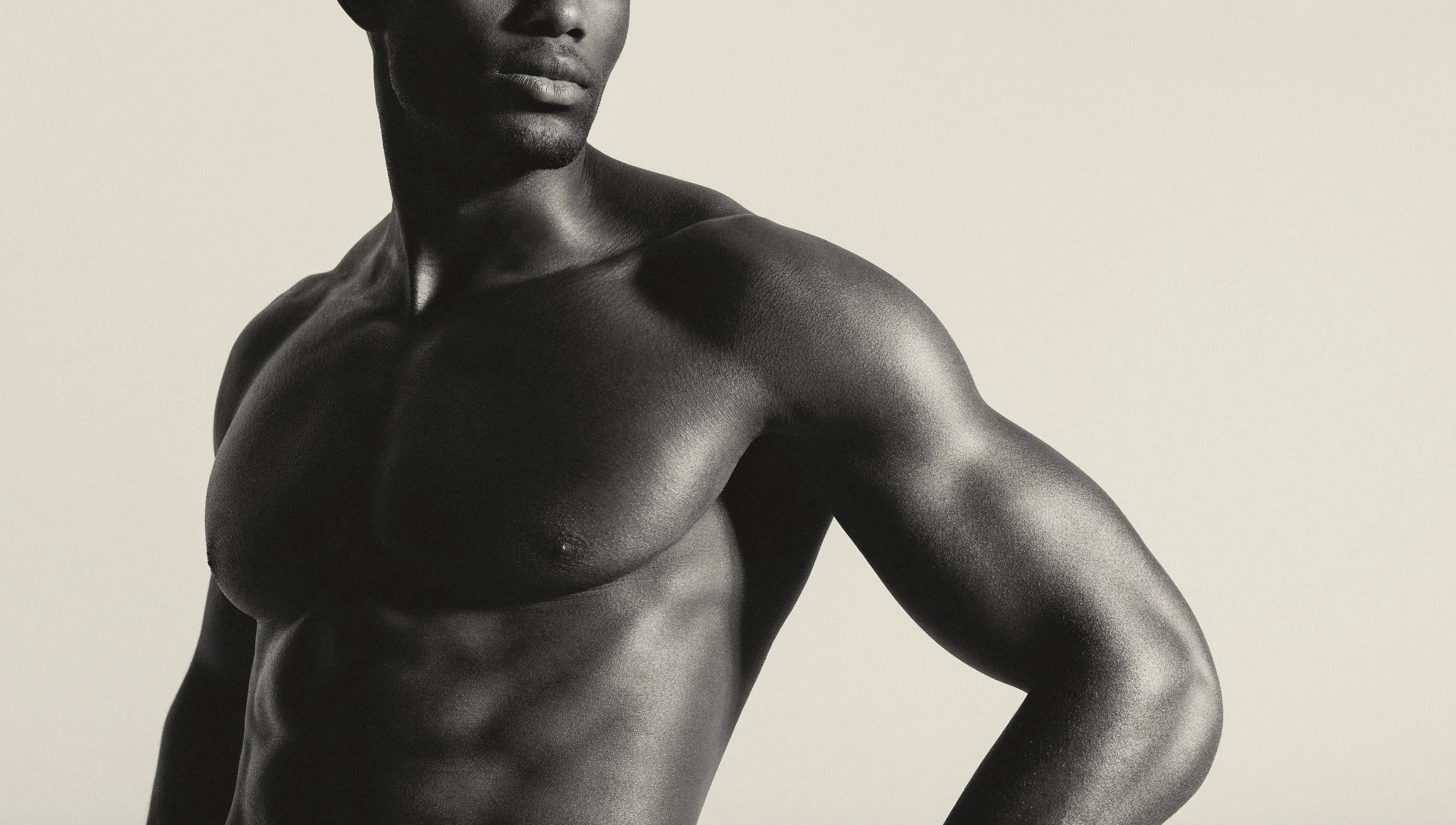 Nahaufnahme eines muskulösen Mannes mit dunkler Haut, der den Oberkörper und einen Teil des Gesichts zeigt, vor hellem Hintergrund.