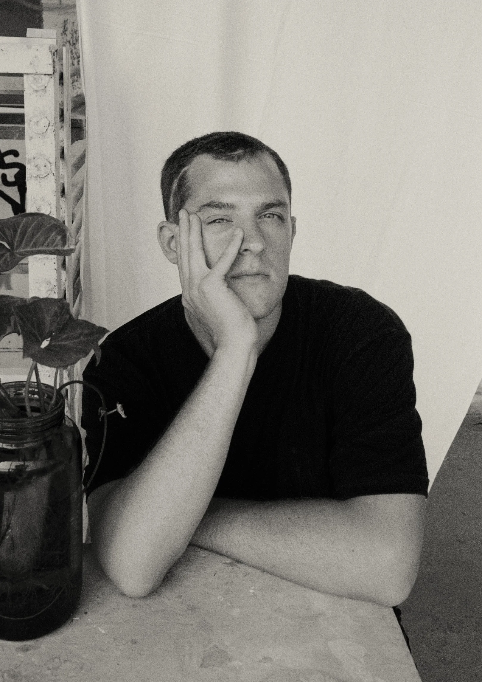 Ein junger Mann sitzt an einem Tisch mit einem Pflanzenglas und sieht in die Kamera.