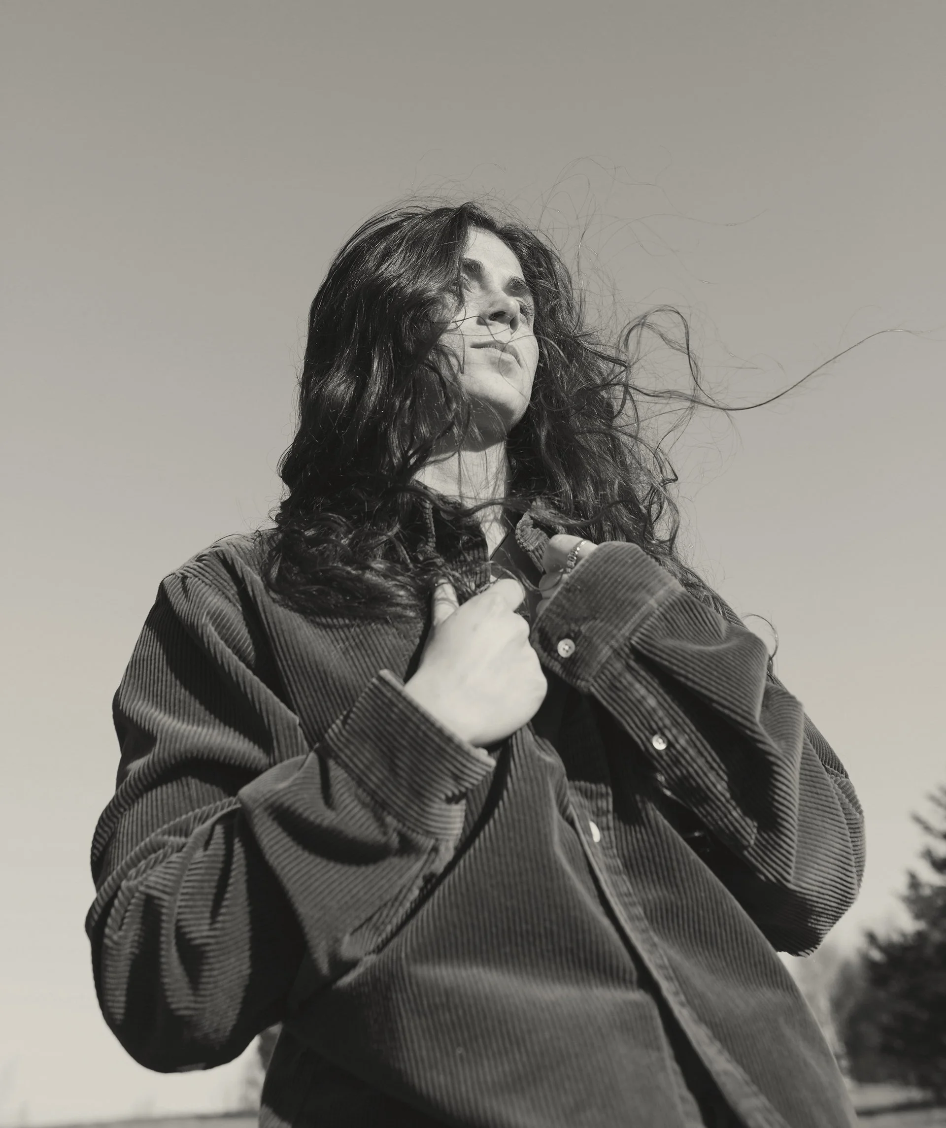 Schwarz-weiß Foto einer jungen Frau mit langen, lockigen Haaren, die nach oben schaut und die Hände an ihrer Jacke hält, im Freien bei klarem Himmel.