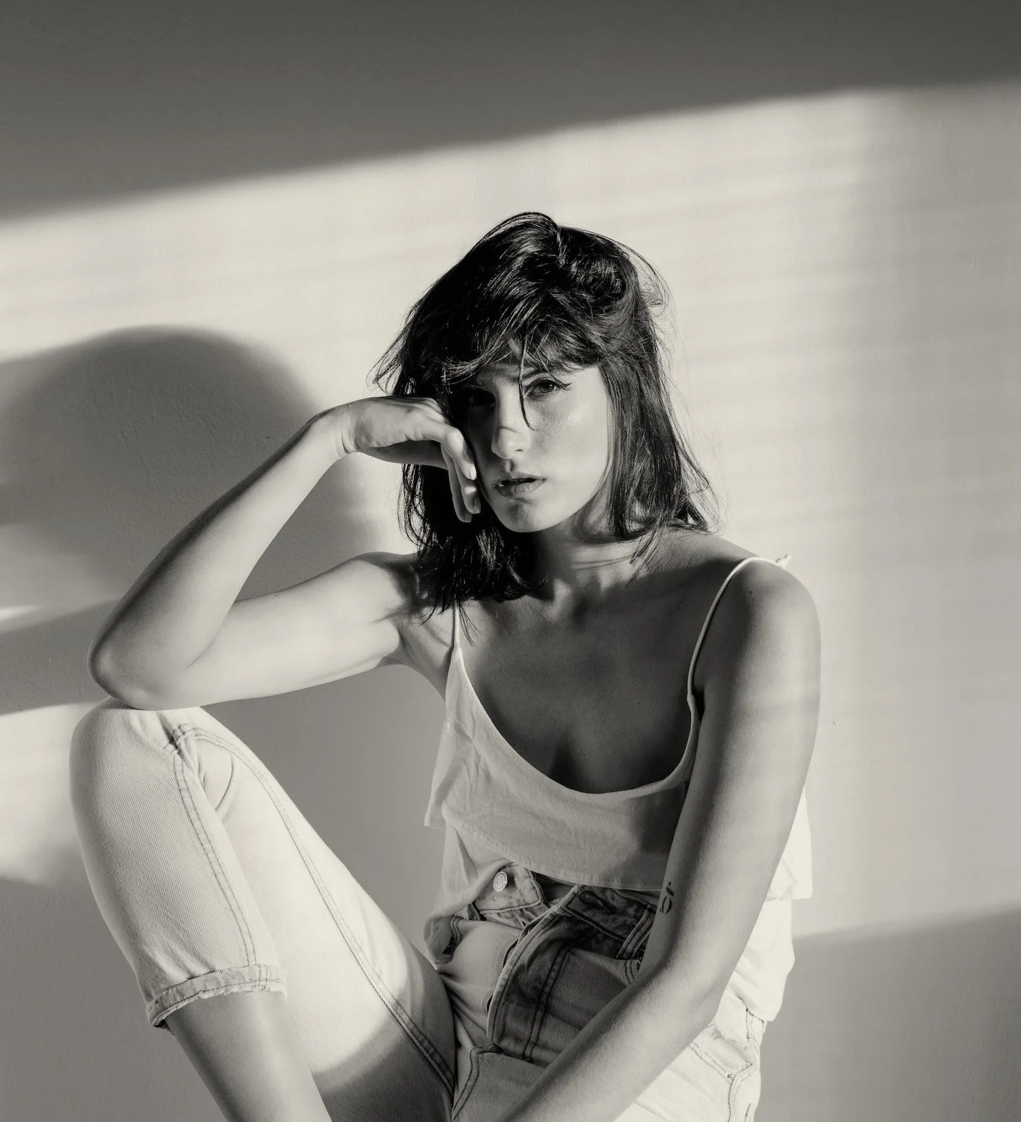 Schwarz-weiß-Foto einer jungen Frau mit mittellangem, gewelltem Haar, die in einem weißen Top und Jeans sitzt, mit Schatten und Licht auf der Wand im Hintergrund.