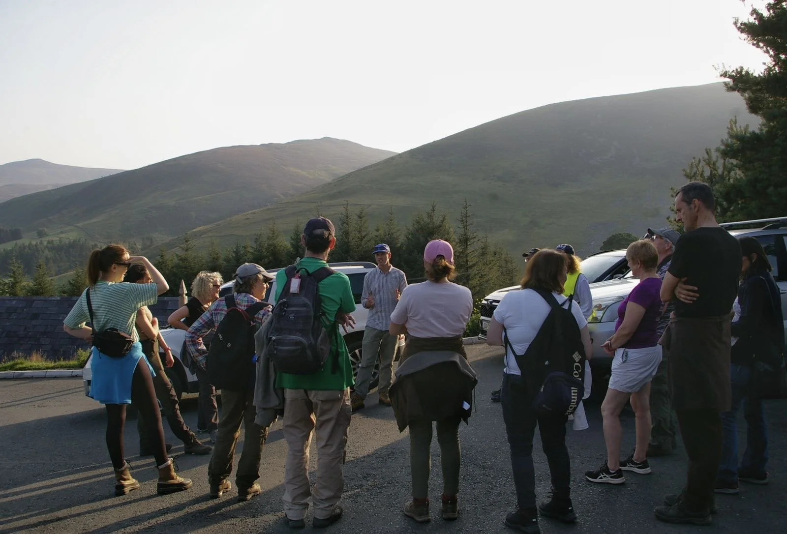 Group of people gathered outdoors on a hillside, with mountains and trees in the background, some listening to a person speaking.