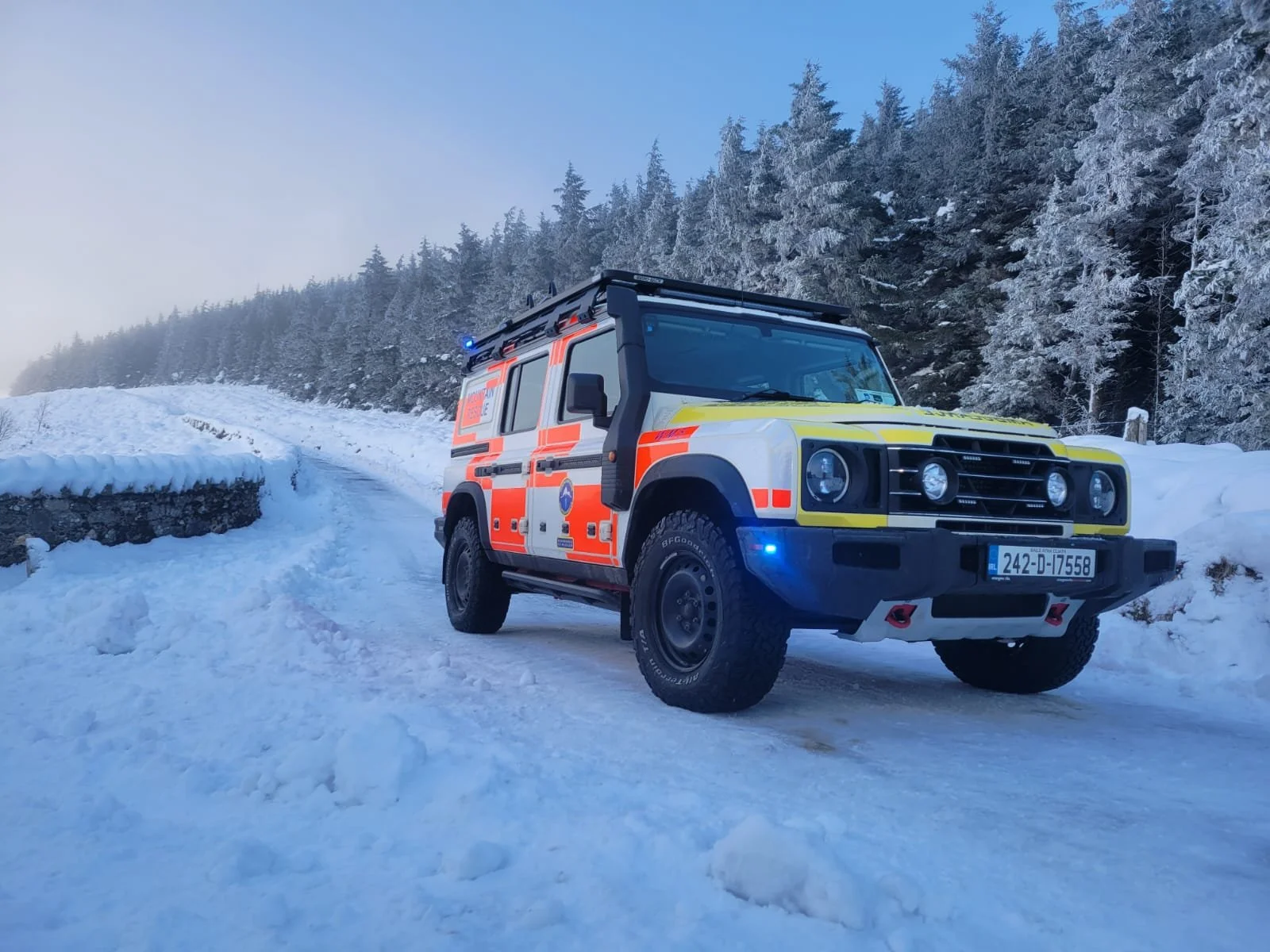 A rescue vehicle on a snow-covered mountain road with snow-covered trees in the background.