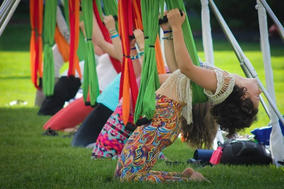 Children doing yoga outdoors in a row with colorful hammocks hanging above them, on a grassy area.