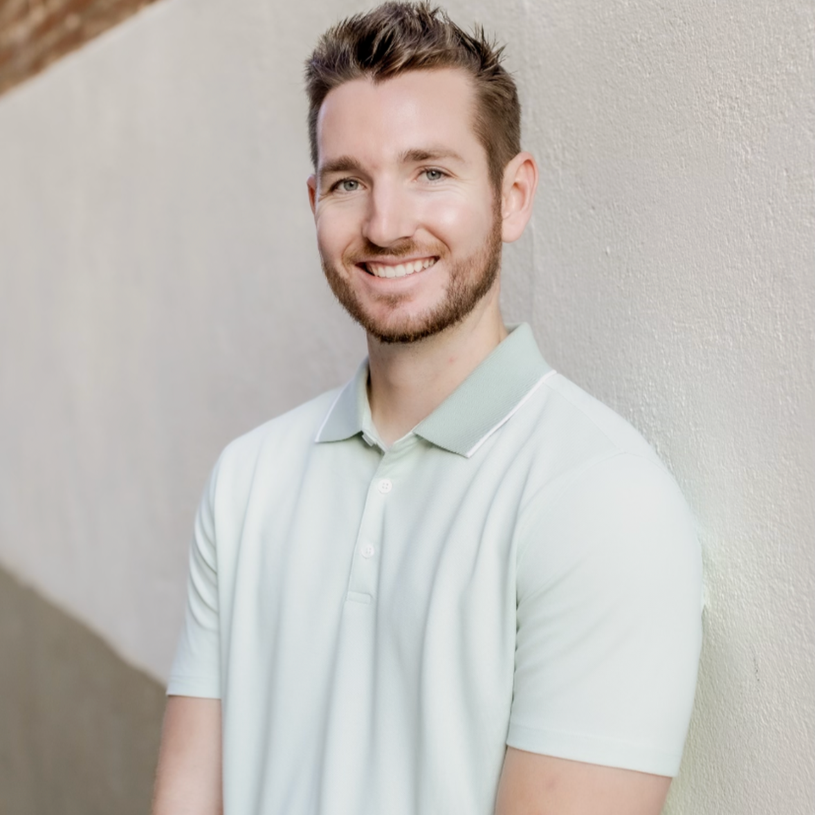 A young man with short brown hair and a beard smiling, wearing a light-colored polo shirt, standing against a light-colored textured wall.
