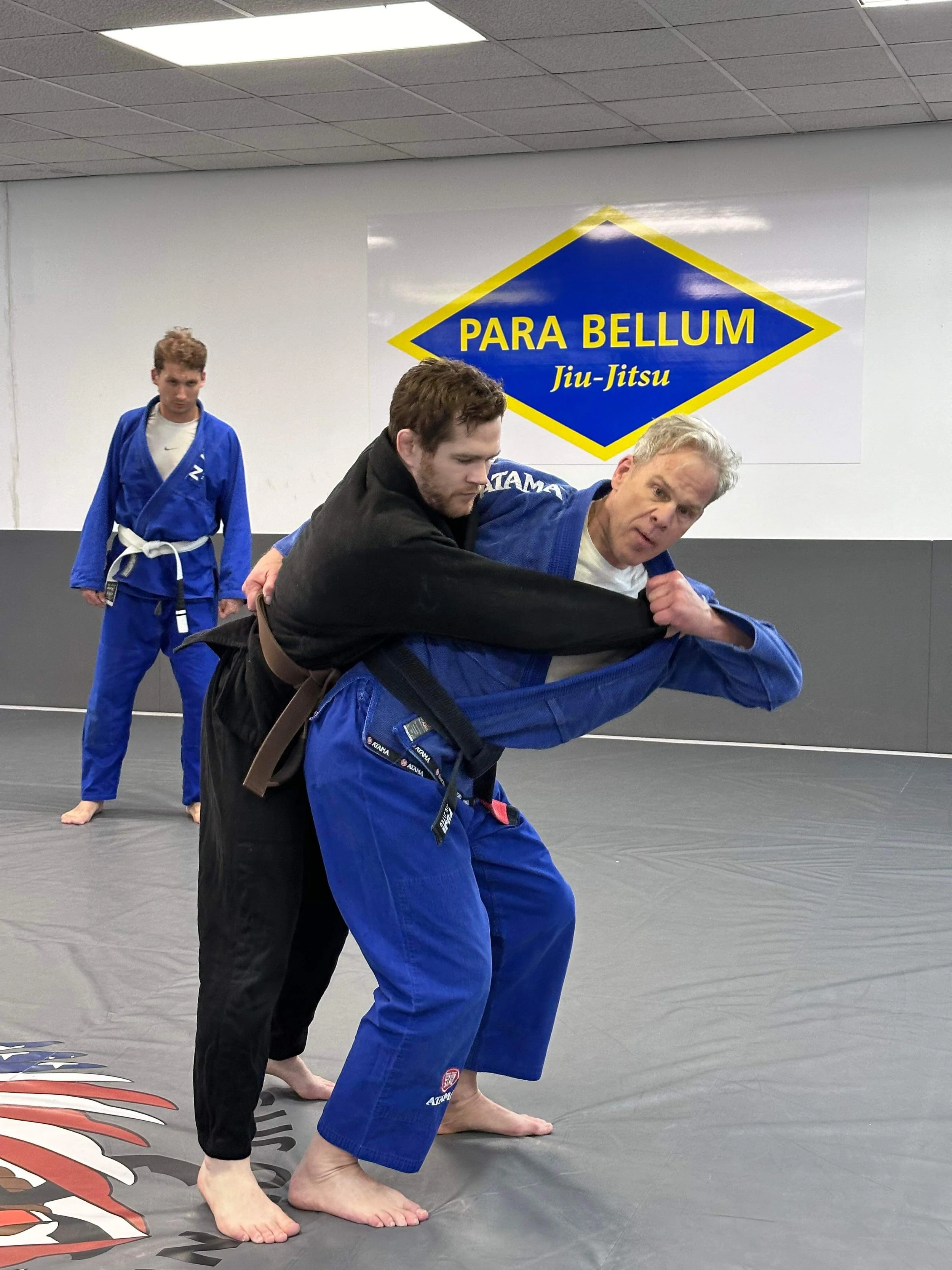 Two men practicing Brazilian Jiu-Jitsu on a mat, with one instructing the other in a grappling move. In the background, a young man in a blue gi observes. A sign on the wall reads 'Para Bellum Jiu-Jitsu'.