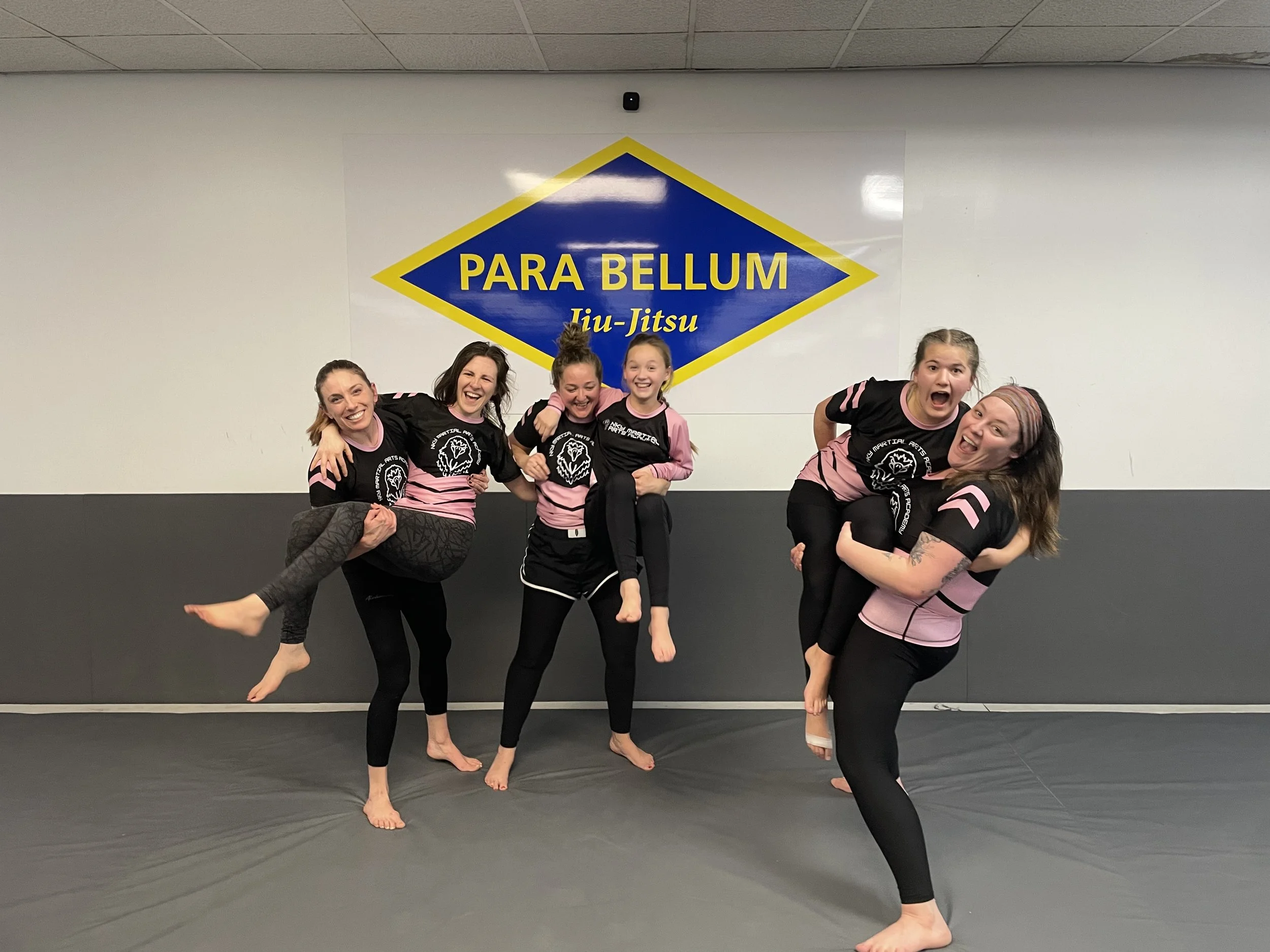 Six women in black and pink martial arts uniforms smiling and posing together in a martial arts studio with a sign that reads "PARA BELLUM Jiu-Jitsu" on the wall behind them.