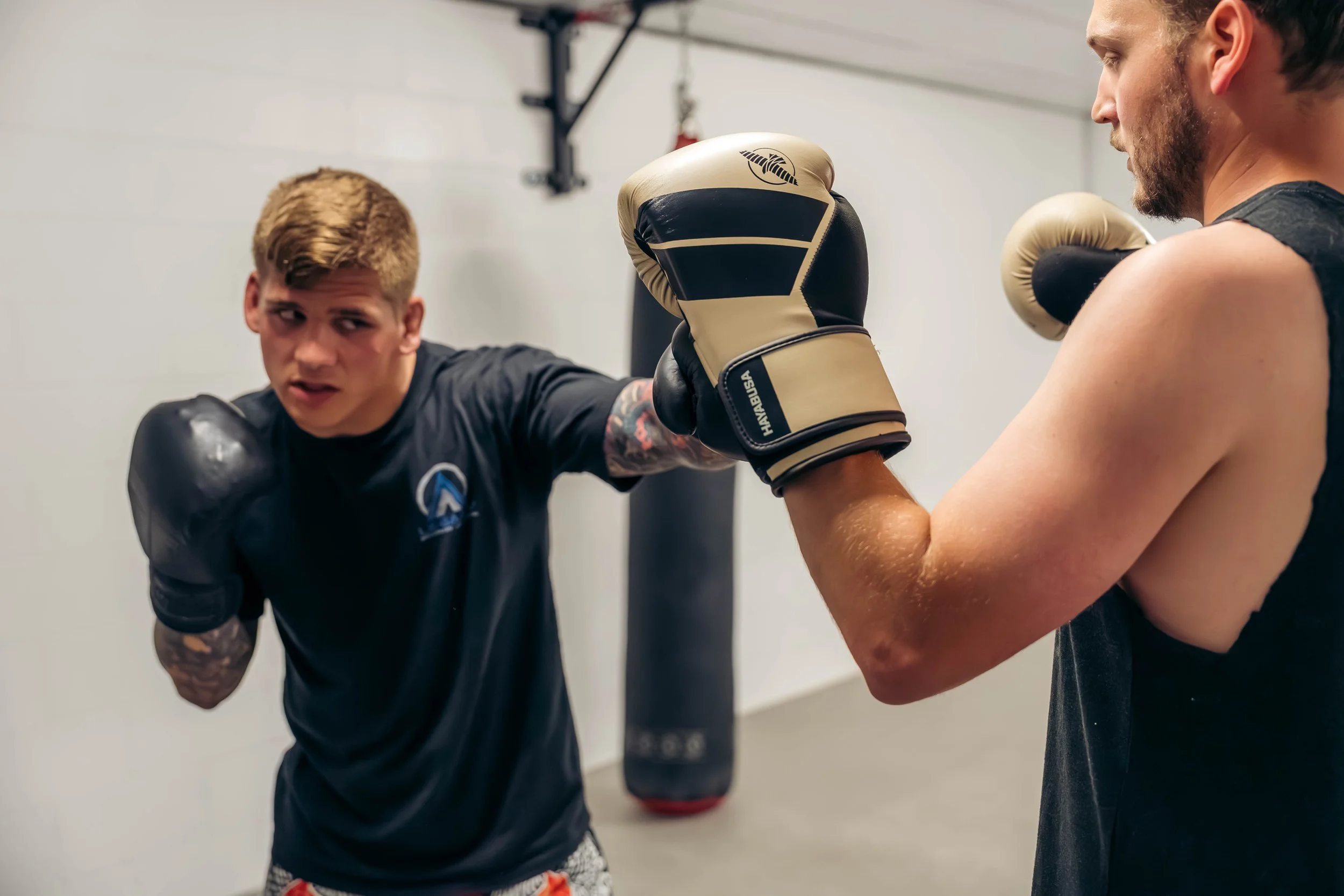 Two men practicing boxing in a gym; one wearing black boxing gloves, the other with beige boxing gloves, with a punching bag in the background.