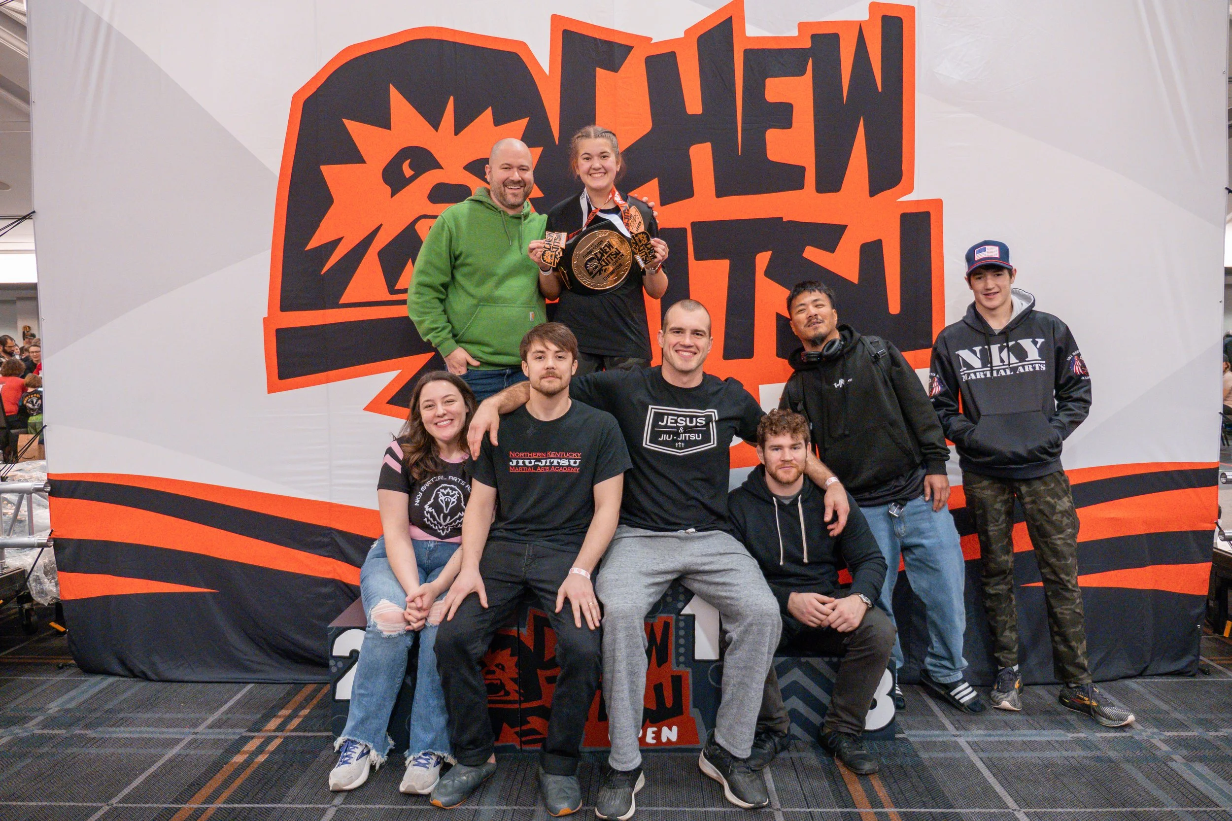 Group of people posing for a photo at a martial arts event with a large 'Nakatani Open' sign in the background. One person is holding a medal and a plaque.