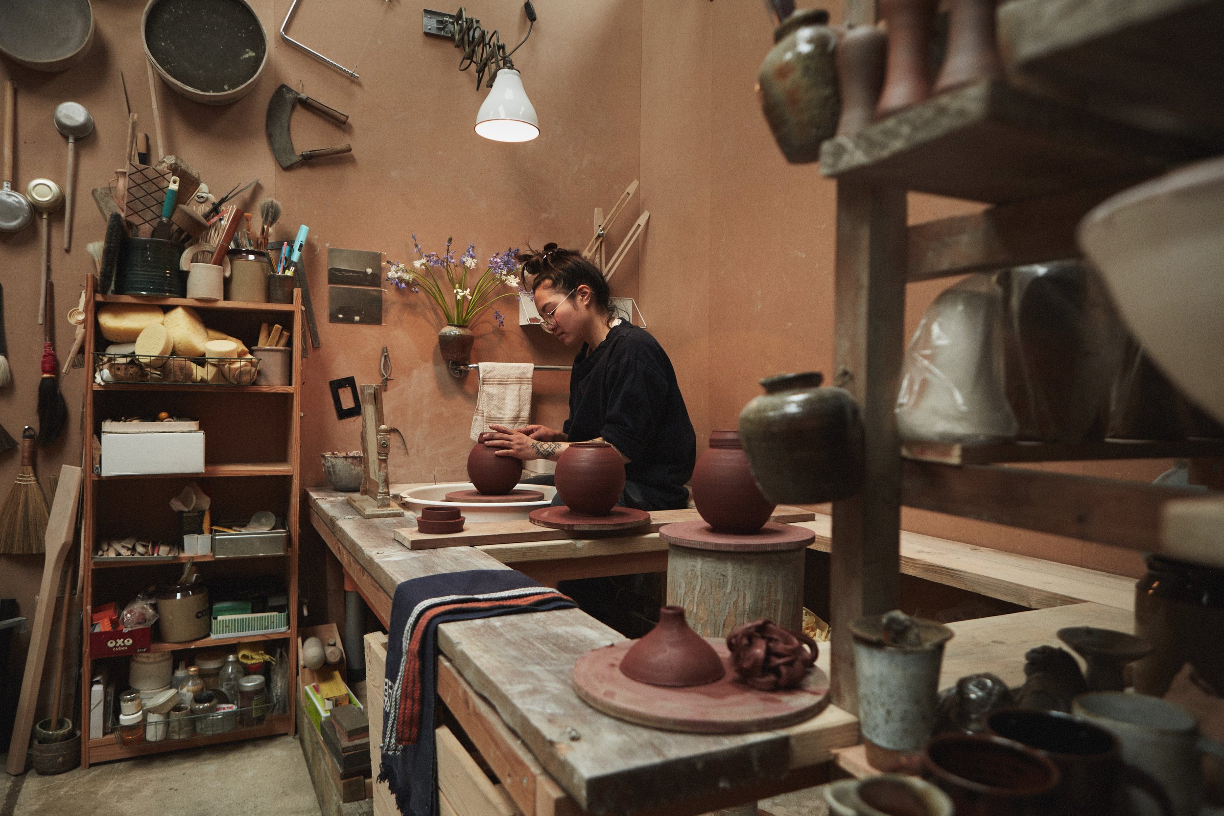 A woman working in a pottery studio, shaping a clay vase on a pottery wheel surrounded by various pottery tools and finished ceramic pieces.