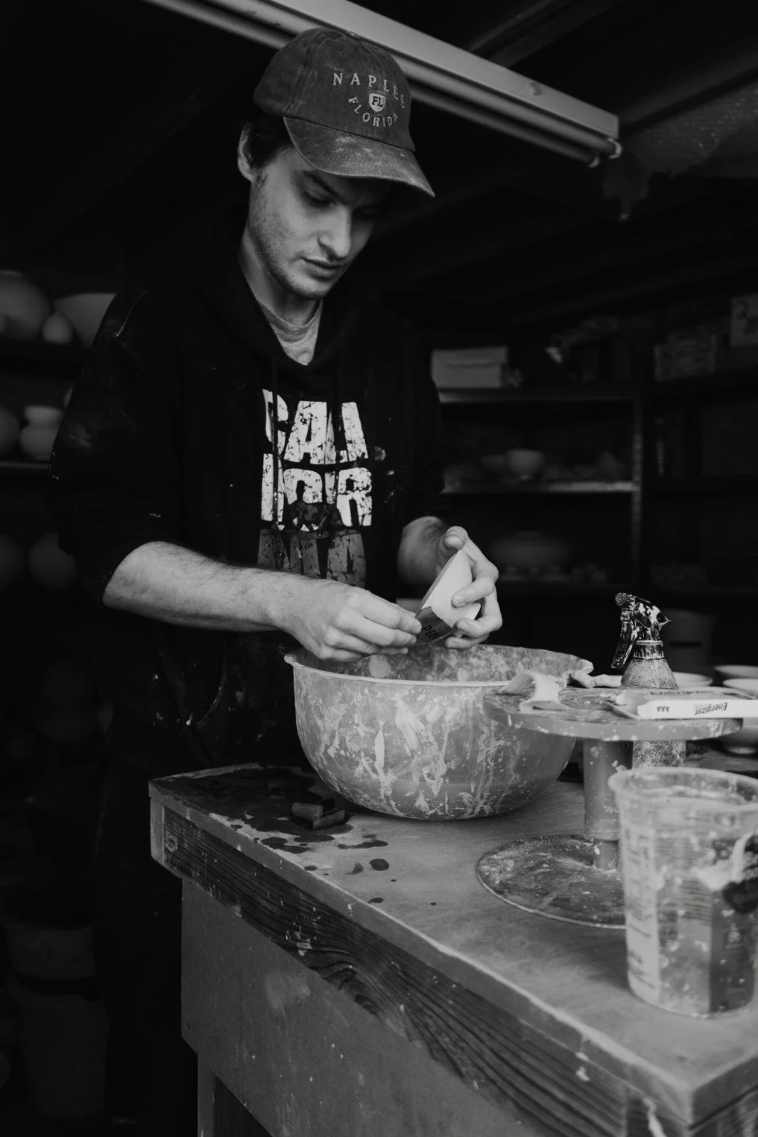 A young man wearing a cap and dark hoodie is working with a bowl and some tools on a wooden workbench, in what appears to be a workshop or studio.