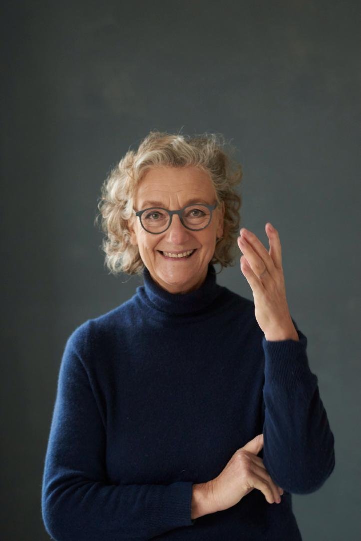 An elderly woman with curly gray hair and glasses, smiling and touching her face with one hand while wearing a navy blue turtleneck sweater, against a plain dark background.