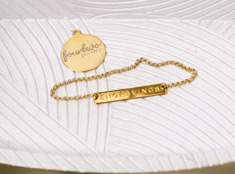 Gold bracelet with two charms, one round with the words 'favourites jewelry' and one rectangular with the word 'ENGLAND' engraved, resting on a white textured surface.
