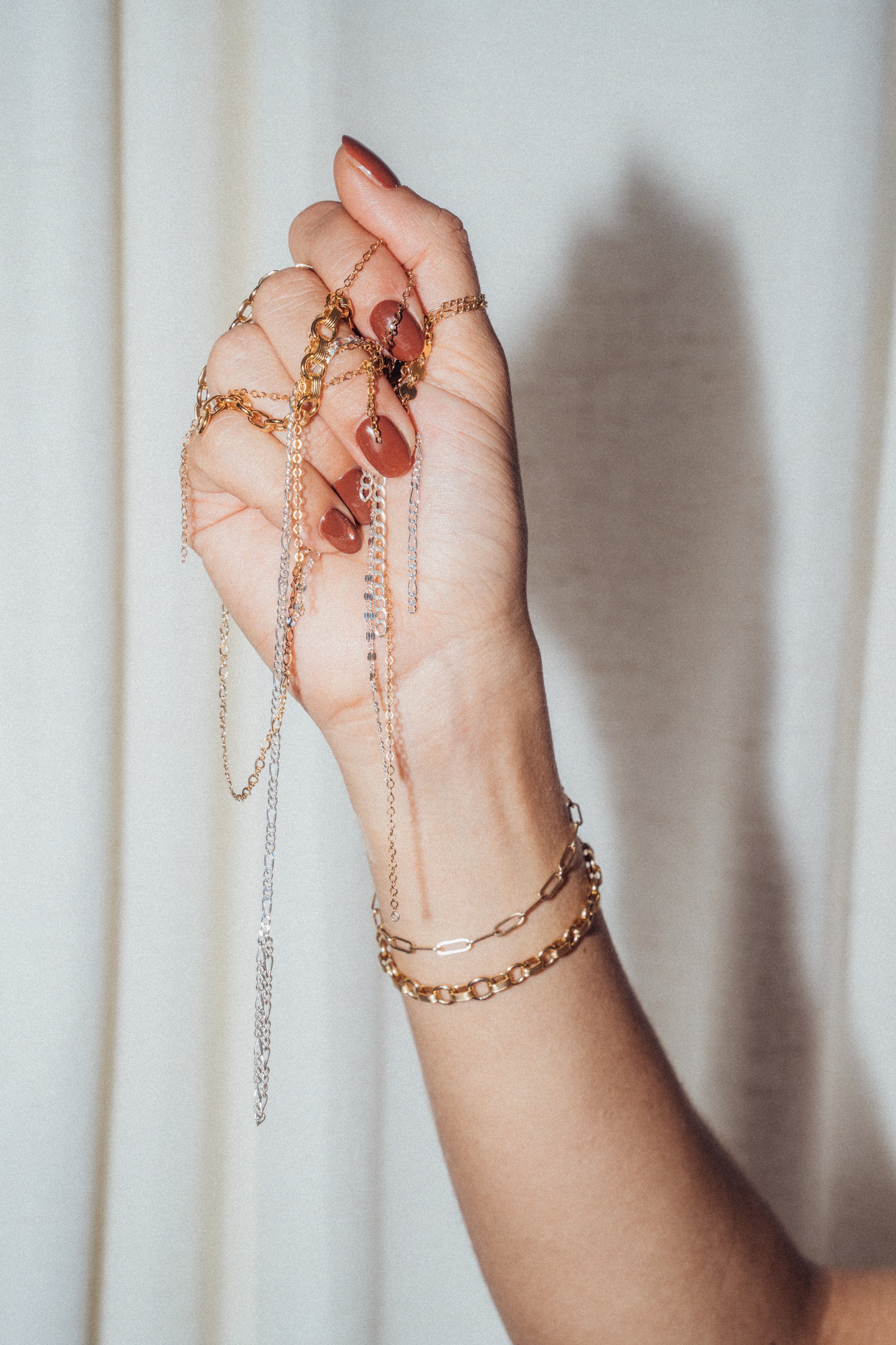 A hand holding multiple gold and silver chain necklaces and bracelets with red painted nails.