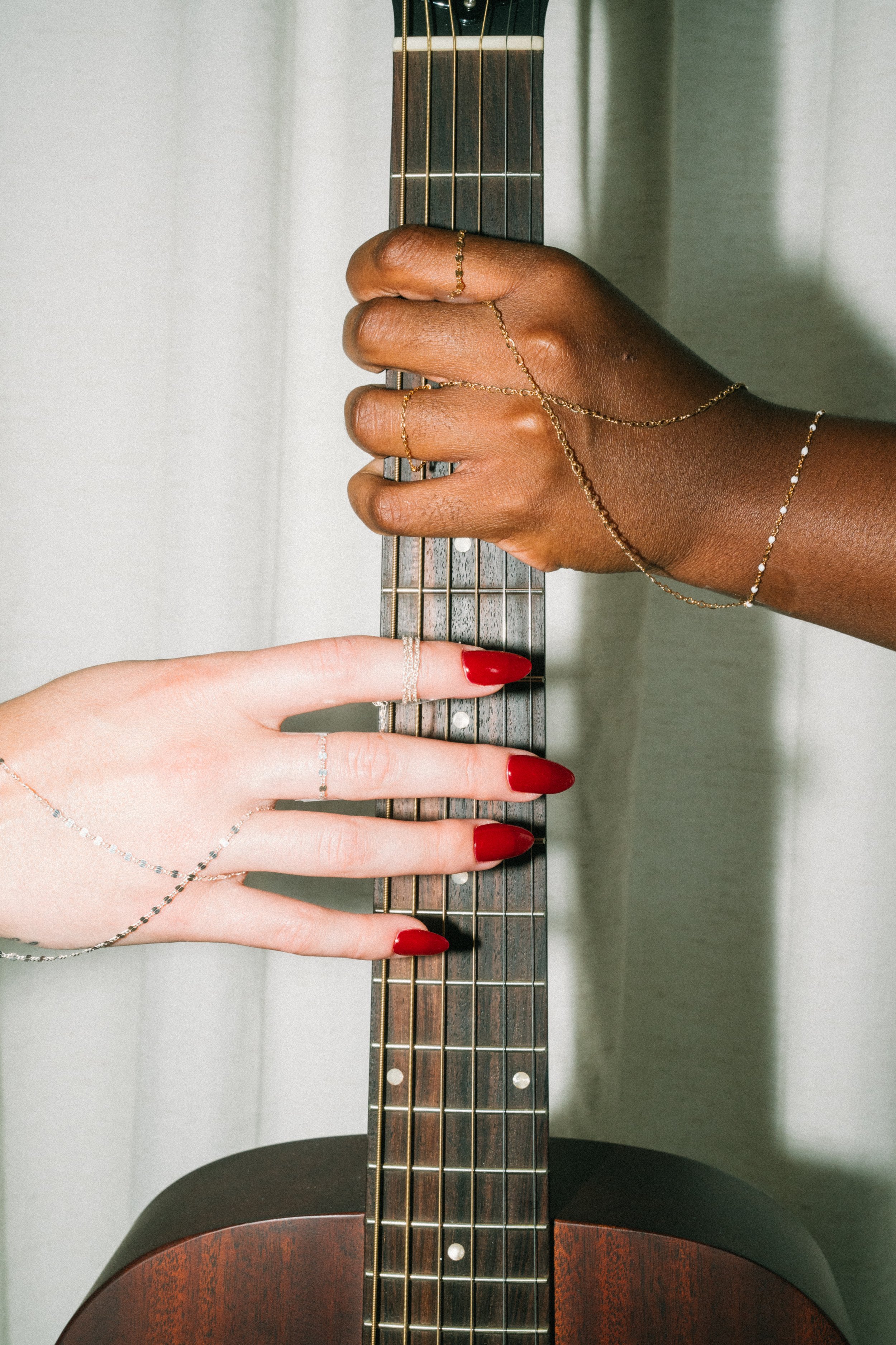 Two hands, one with dark skin and one with light skin, gently touching a guitar neck. Both hands are adorned with jewelry, including rings, bracelets, and chains, and have red manicure.