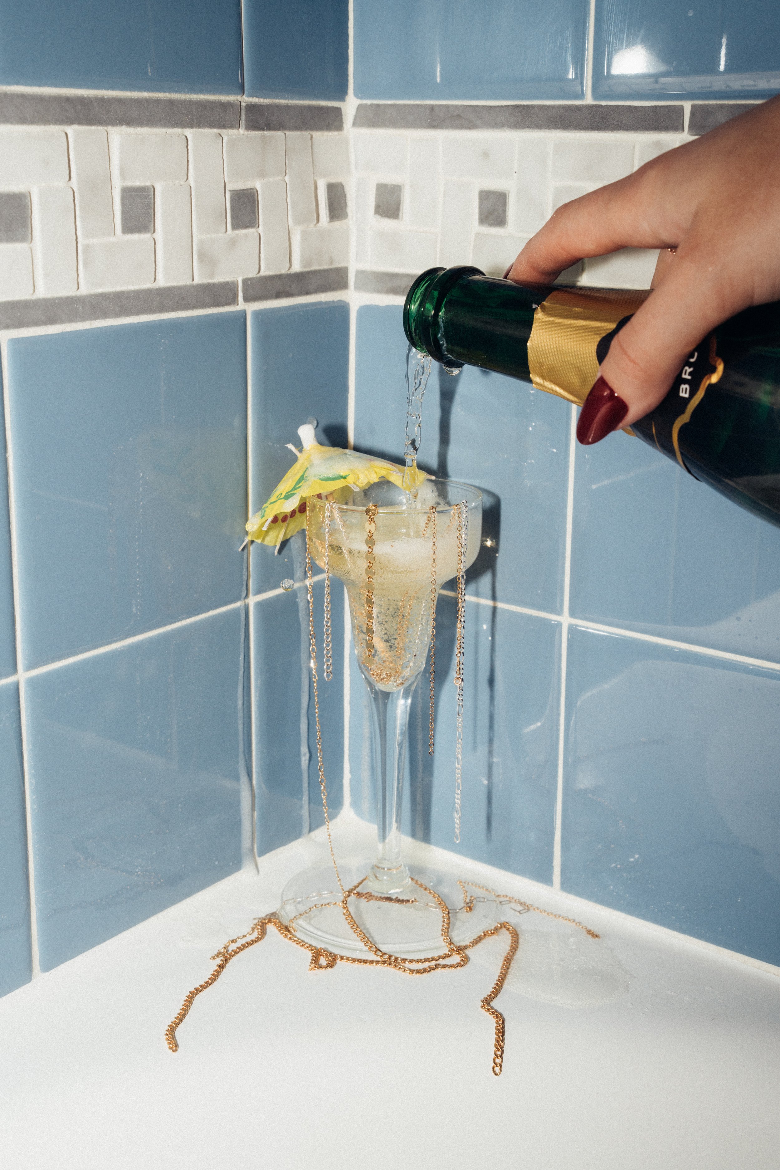 A hand pouring champagne into a glass shaped like a martini glass with a mini umbrella, decorated with gold chains, sitting on a white surface against blue and grey tiled wall.