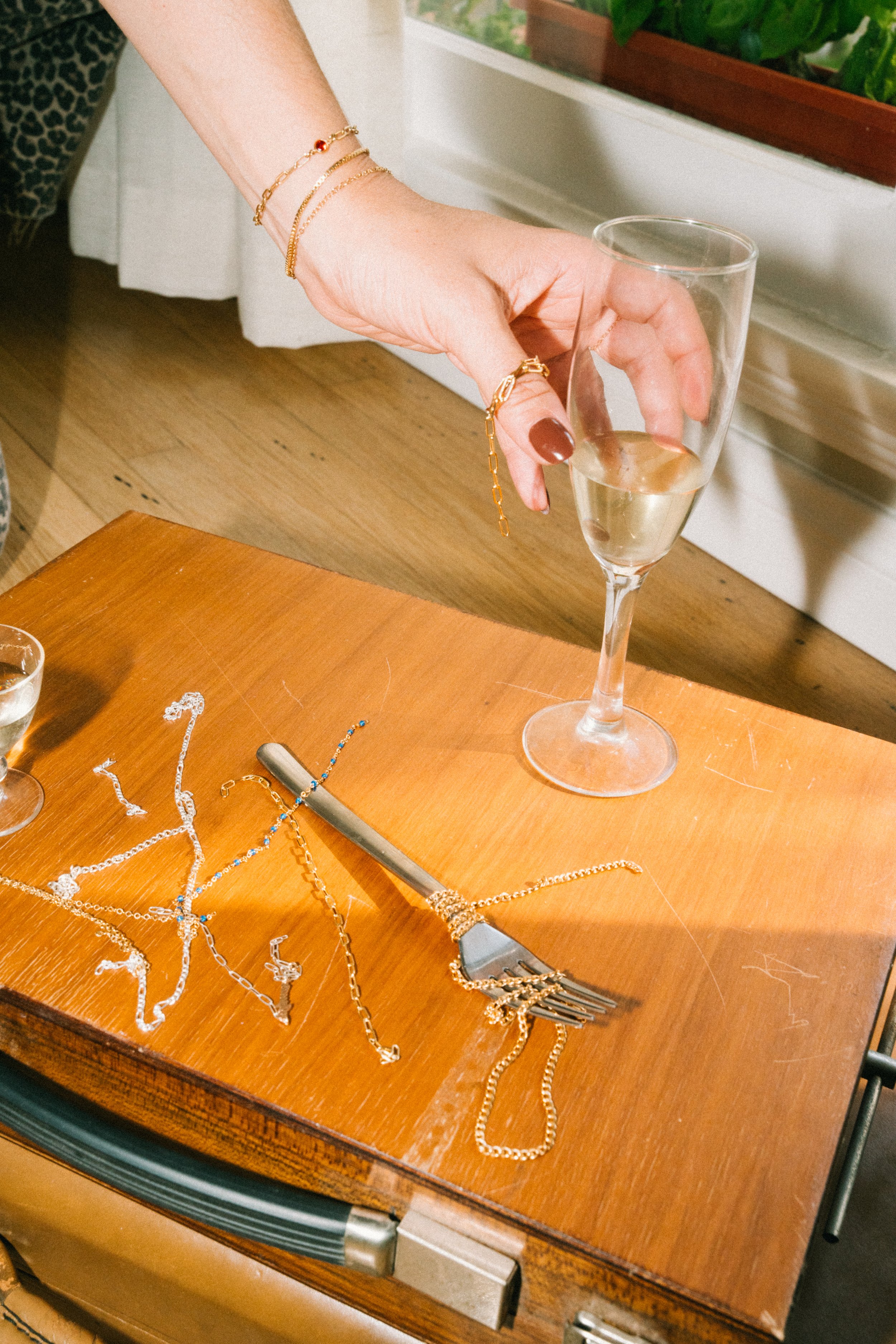 A woman places a glass of champagne on a wooden table scattered with jewelry and a fork.