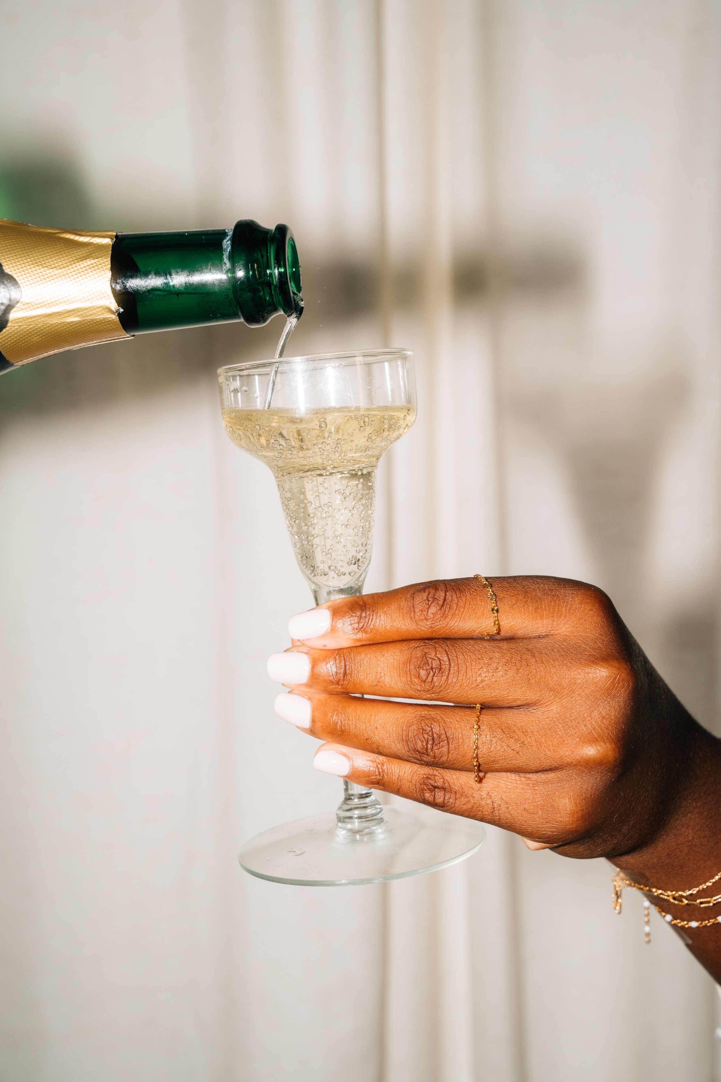 Person pouring champagne into a flute glass, with a hand holding the glass, adorned with rings and delicate jewelry.