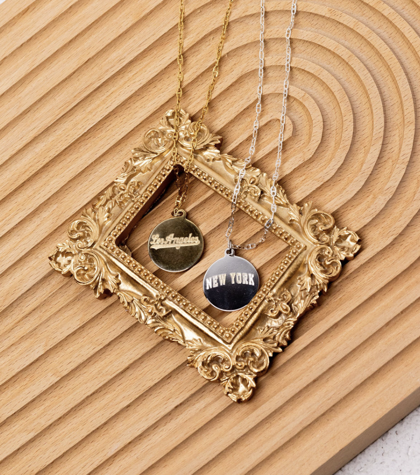 Two necklaces, one gold and one silver, are hanging over an ornate gold picture frame lying on a wooden surface. The gold necklace has a round pendant that says "Los Angeles" and the silver necklace has a round pendant that says "New York."