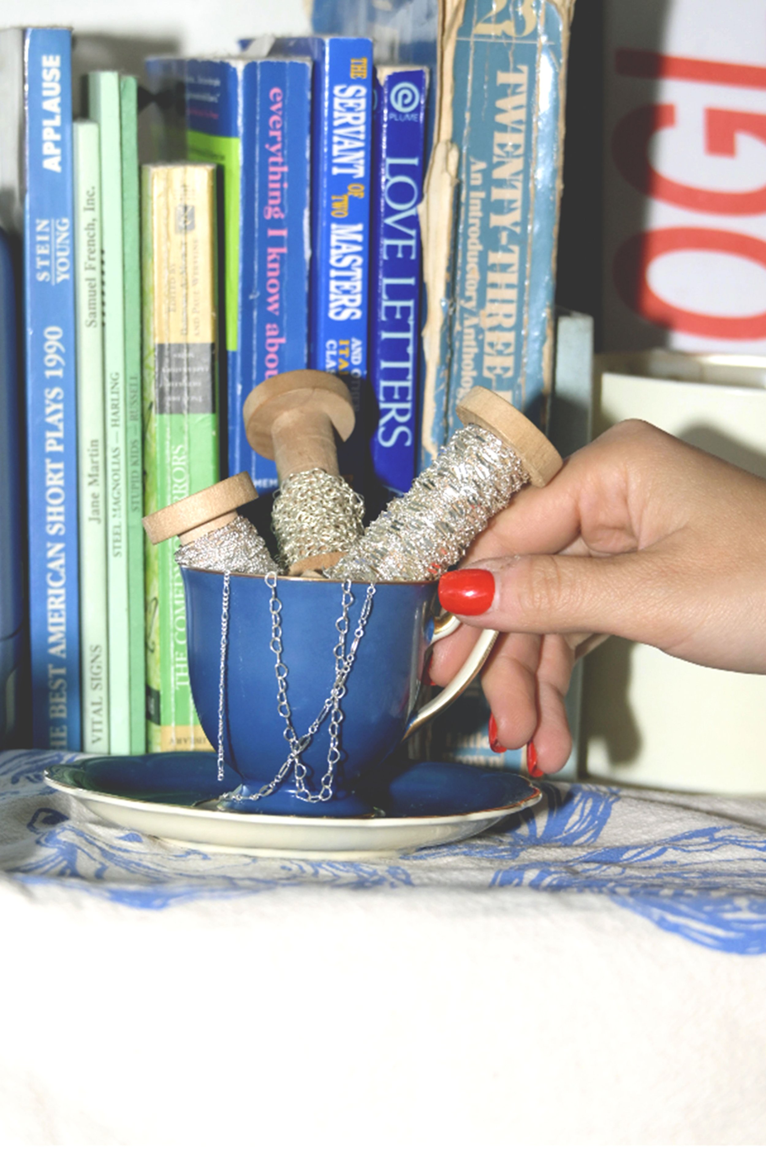 A hand with red painted nails holding a blue teacup filled with silver jewelry, surrounded by books on a shelf, with a saucer underneath on a table with a printed cloth.