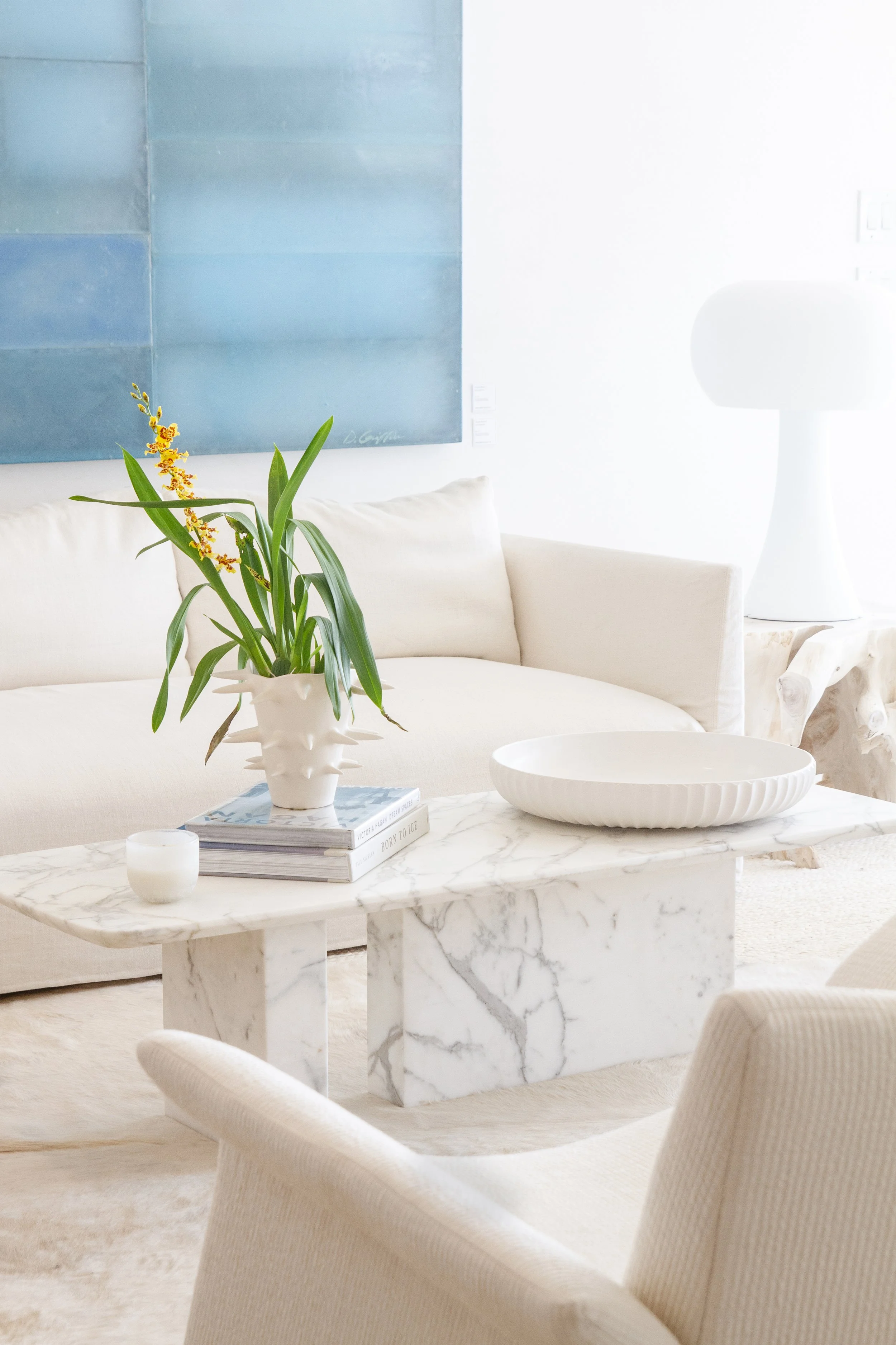 A bright, minimalist living room featuring a white sofa, a white marble coffee table with a decorative bowl, a stack of books, a white vase with green plants and yellow flowers, and a large blue painting on the wall.