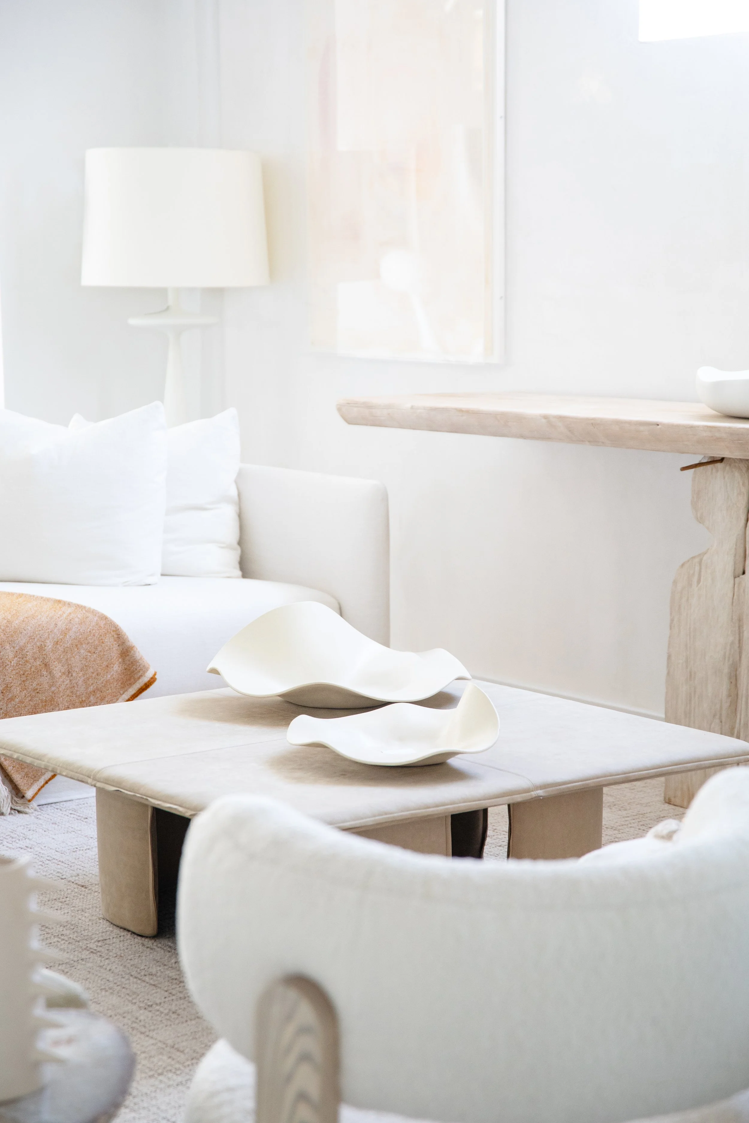 Minimalist living room with white sofa, cream armchair, wooden coffee table with abstract white bowls, and soft neutral decor.